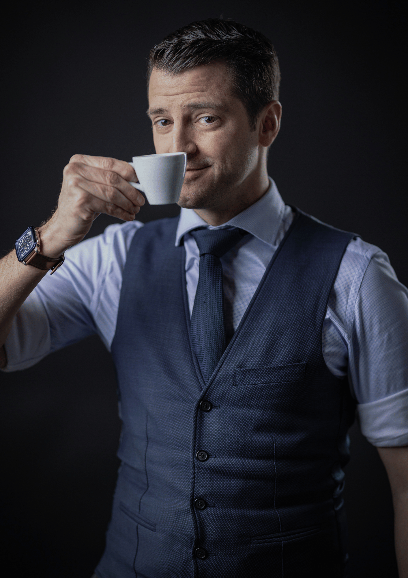 A man in a business suit drinking coffee against a dark background.