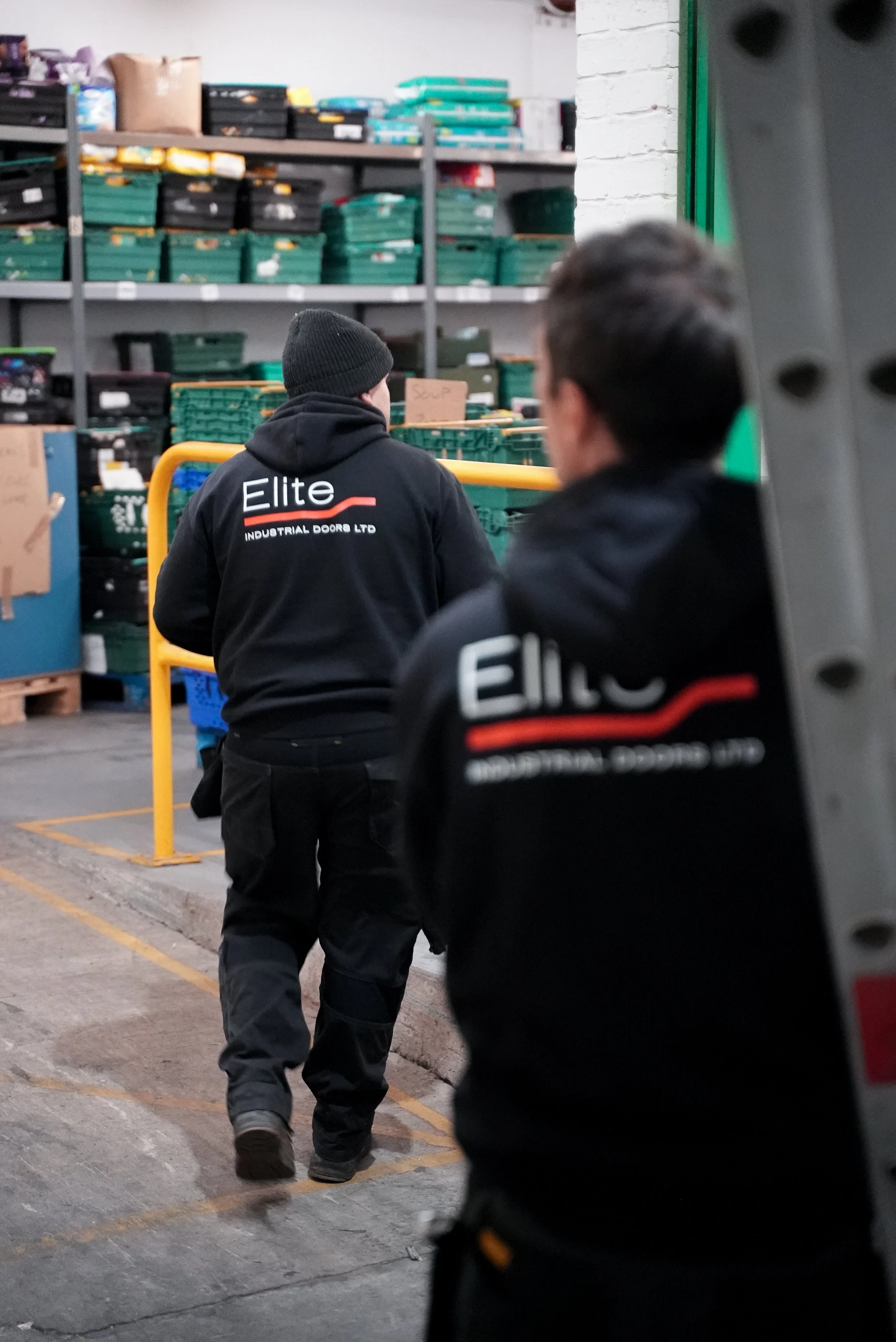 Two workers in black hoodies with 'Elite Industrial Doors Ltd' logo working in a warehouse with shelves of boxes and crates.