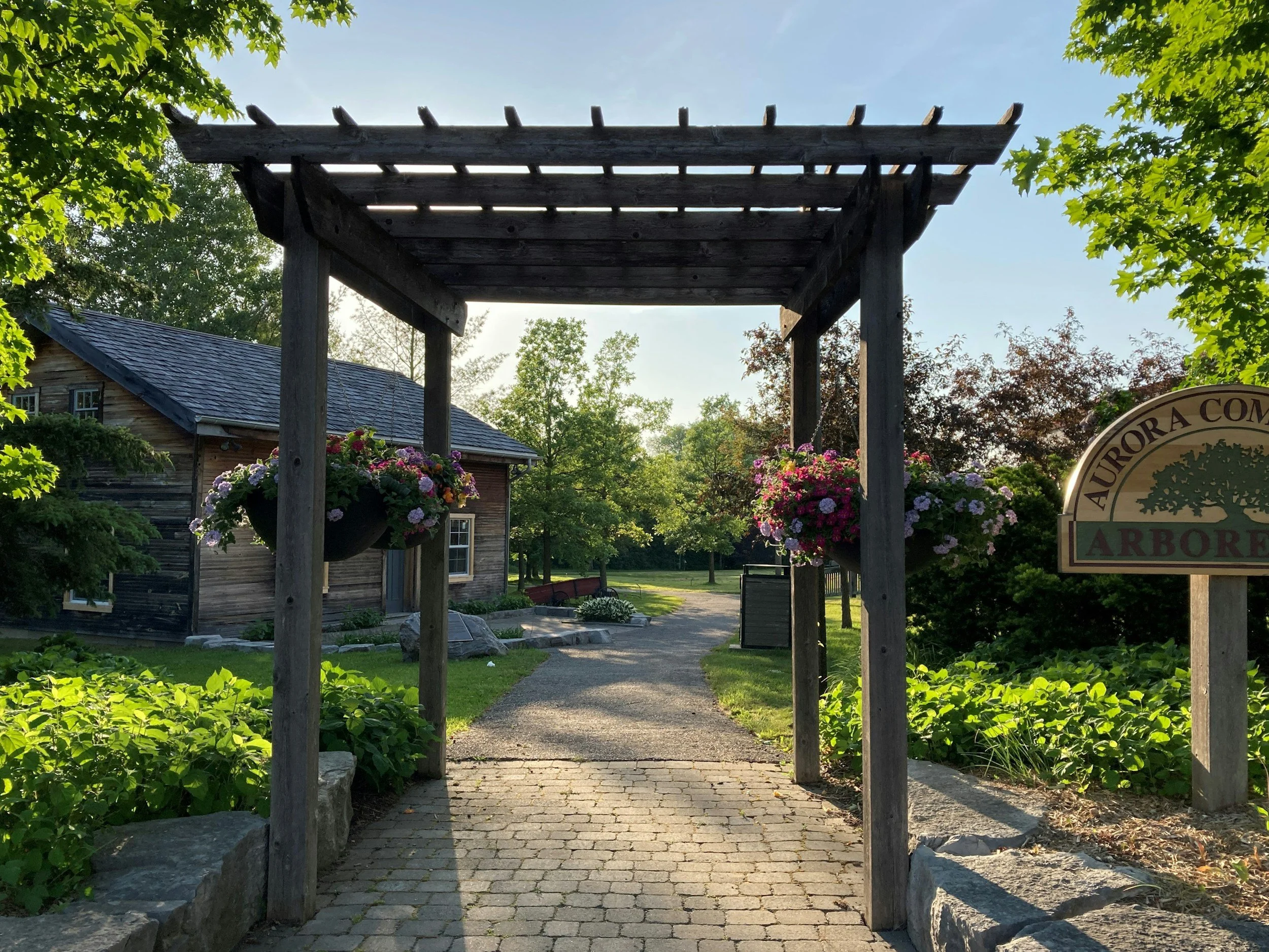Pathway through a wooden archway with hanging flower baskets, leading to a garden area with trees and a building on the left, with a sign reading "Aurora County Arboretum."