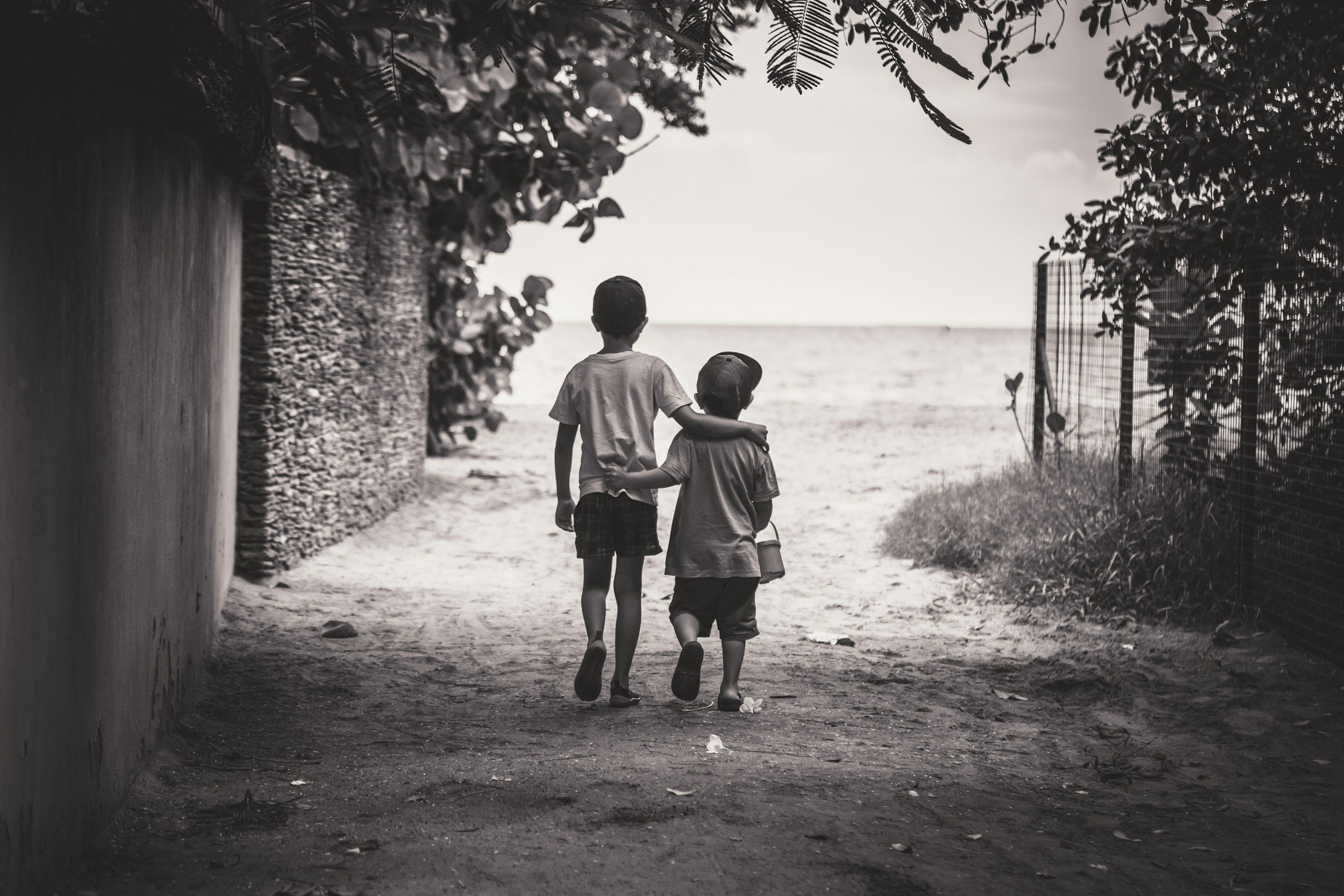 Two children walking towards the beach on a sandy path, with the ocean visible in the background, one with arm around the other's shoulders.