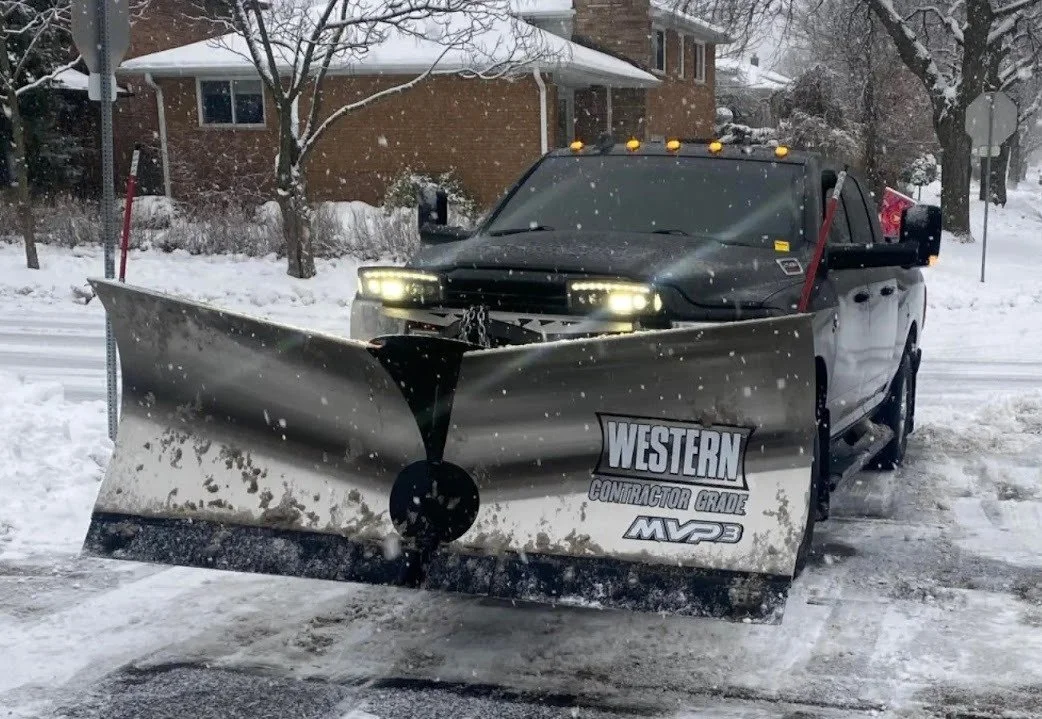 A black pickup truck equipped with a Western Convector Gblade snow plow, clearing snow from the street in a residential area during winter.