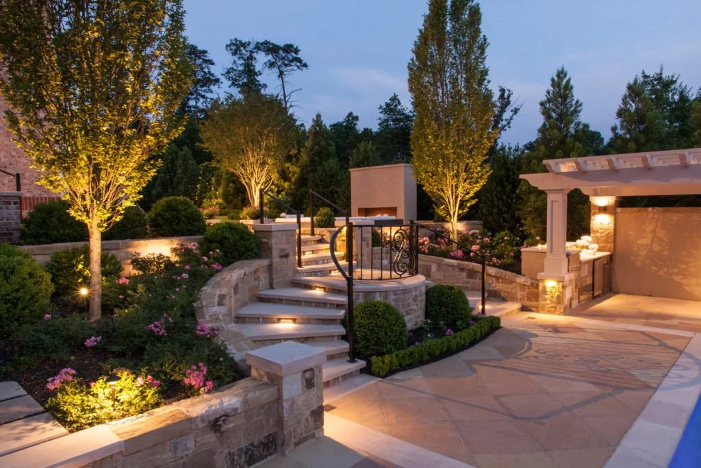 A landscaped backyard patio at dusk with illuminated trees, flowering bushes, stone stairs, and a seating area.