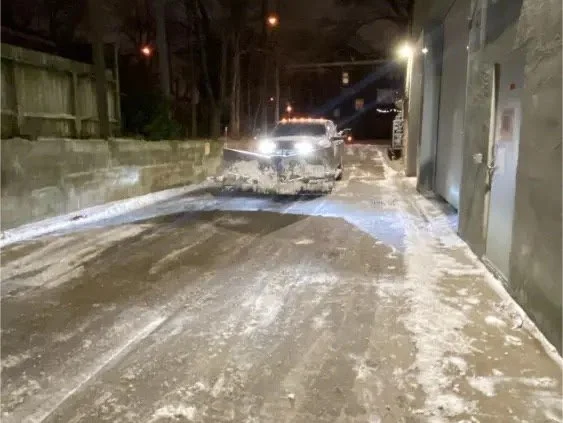 A snow-covered city street with a damaged car parked along the sidewalk at night.
