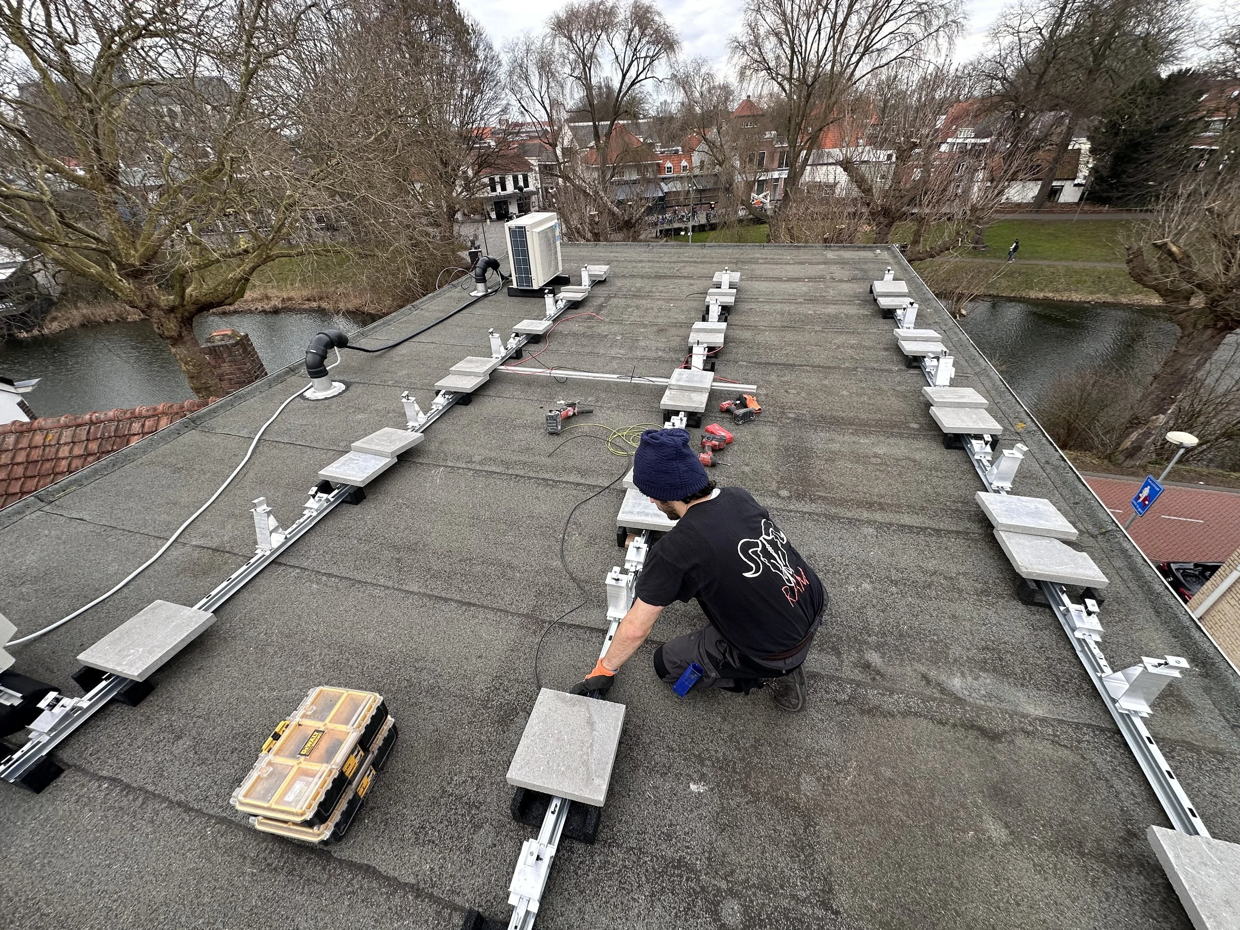 Een man op een plat dak met zonnepanelen, hij werkt met gereedschap, omringd door elektrische kabels en apparatuur, met een rivieroever en bomen op de achtergrond.