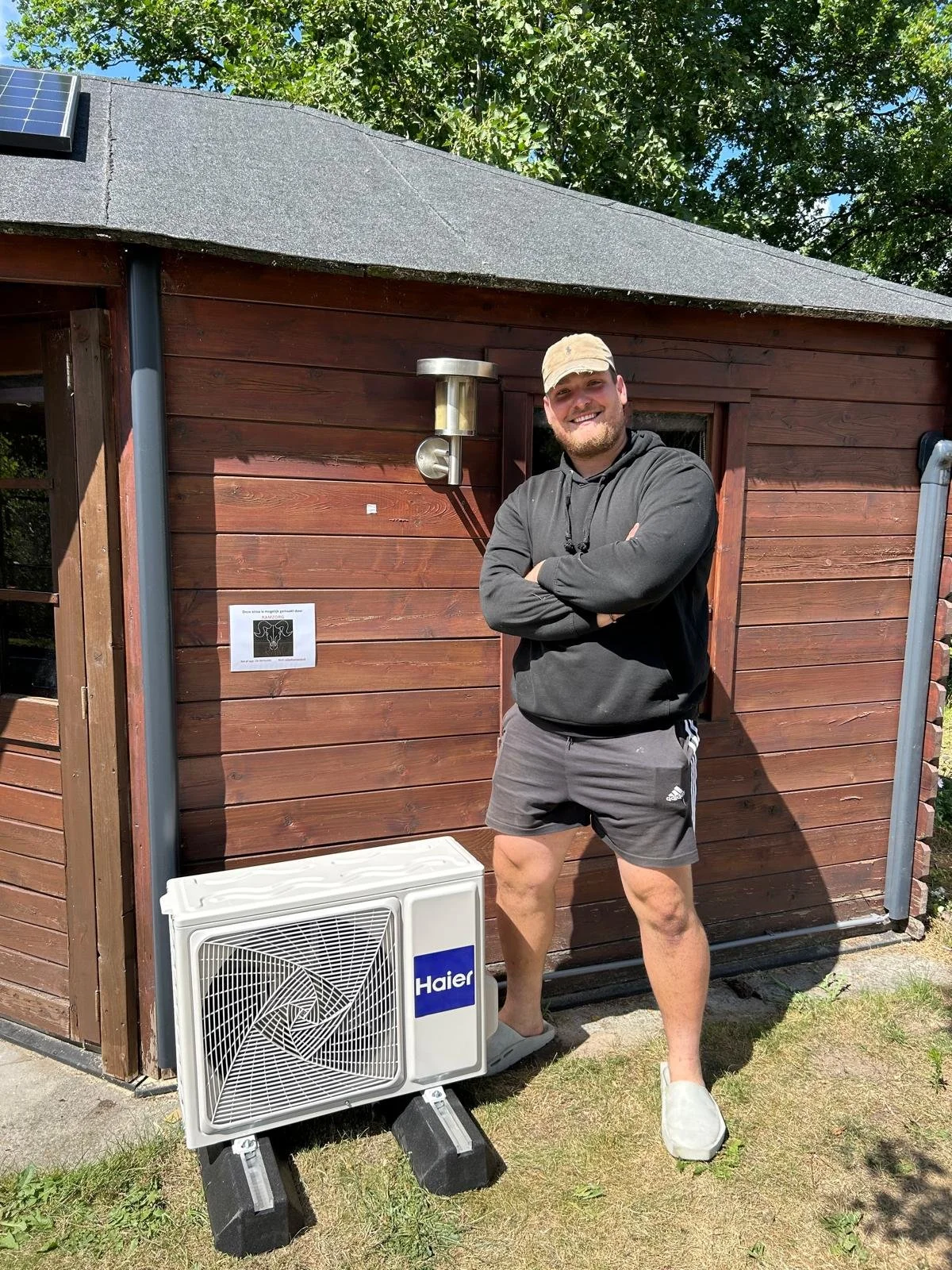 Man staat buiten voor een houten bijgebouw, gekleed in een zwarte hoodie, korte broek en slippers, met naast hem een buitenunit van een airconditioner van het merk Haier.