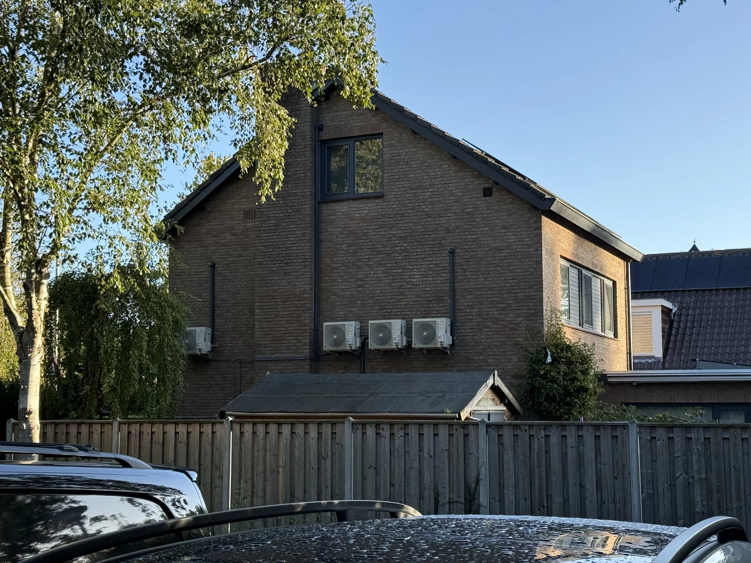 Een huis met een bakstenen gevel, meerdere airconditionings units aan de muur, een schutting en een boom voor het huis, onder een heldere blauwe hemel.