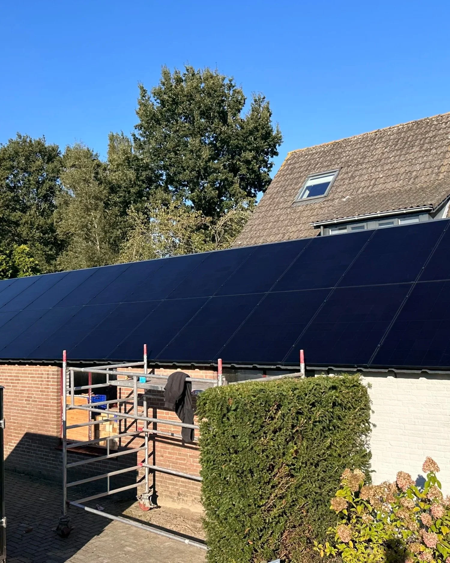 Zwarte zonnepanelen op het dak van een huis met een dakkapel, omgeven door bomen en een heg, onder een blauwe hemel.
