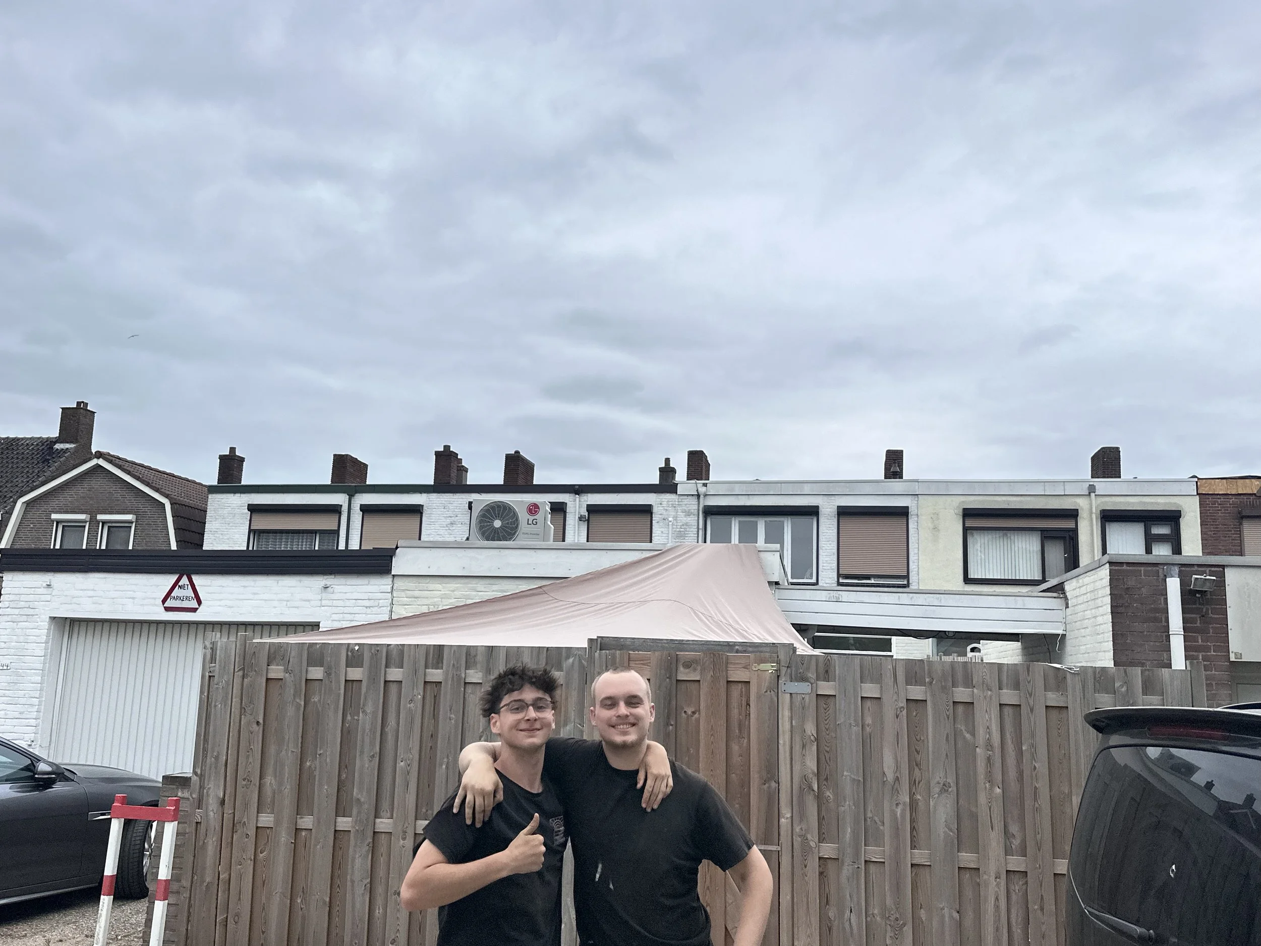 Twee jongens staan samen buiten, armen om elkaar, voor een houten hek met een parkeerplaats en appartementen op de achtergrond. Het is bewolkt.