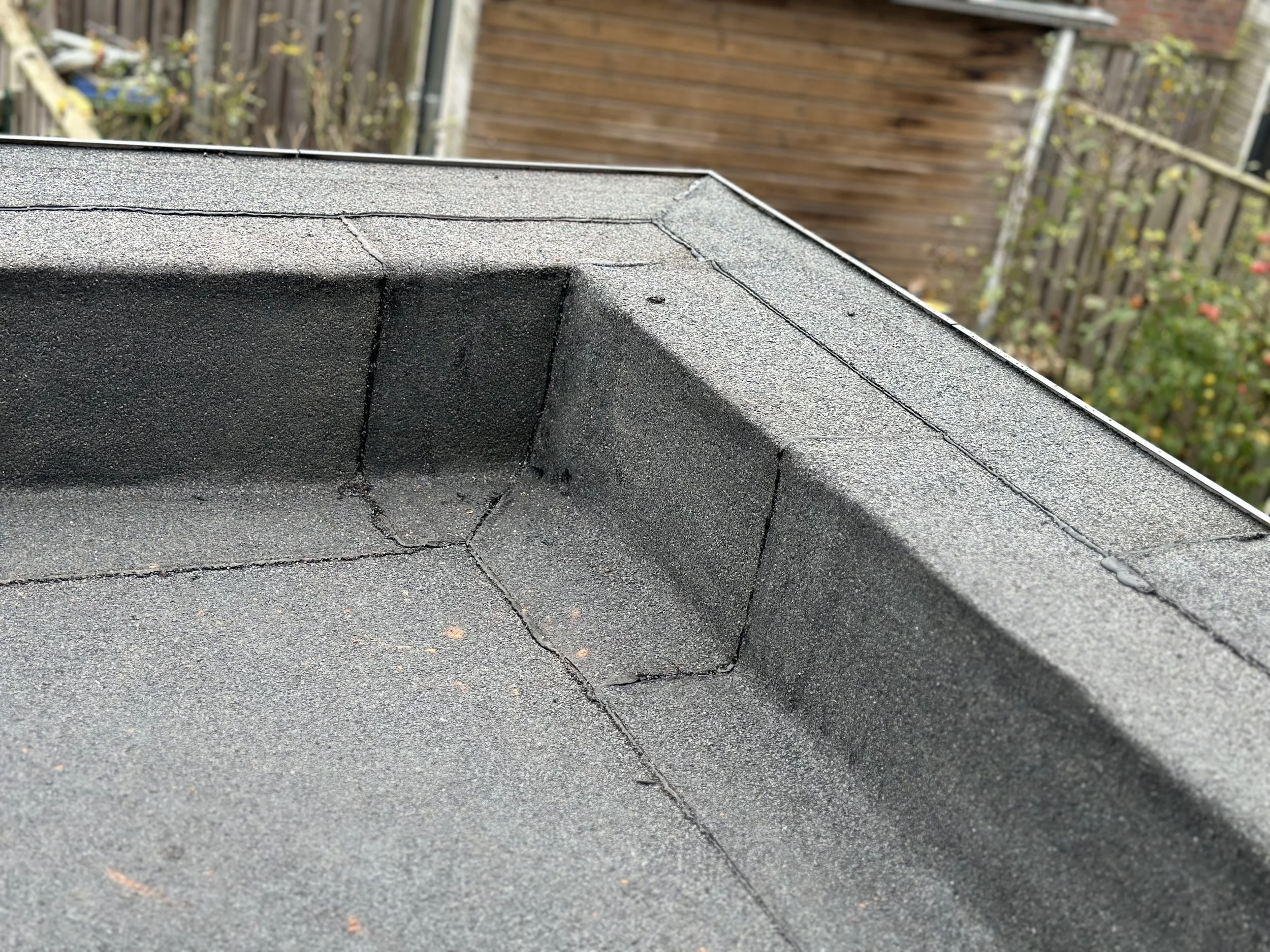 Close-up van een dak gemaakt van bitumen shingles, zichtbaar net gerenoveerd, in een tuin met houten hekken en planten op de achtergrond.