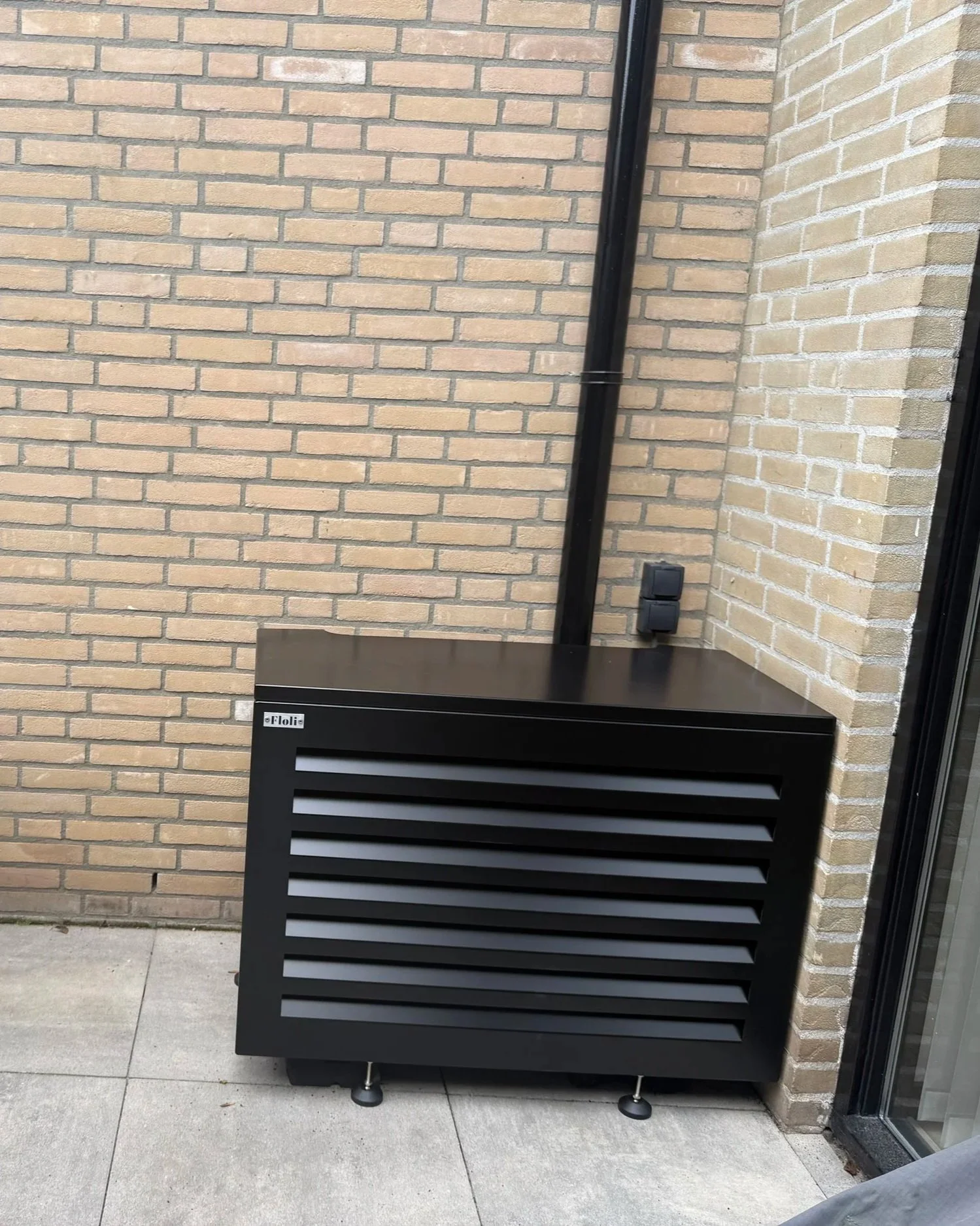 Een zwart ventilatiekast met horizontale ventilatieroosters, merk 'Floli', staat buiten tegen een bakstenen muur, grijs tegelvloer en een glasdeur zichtbaar rechts.