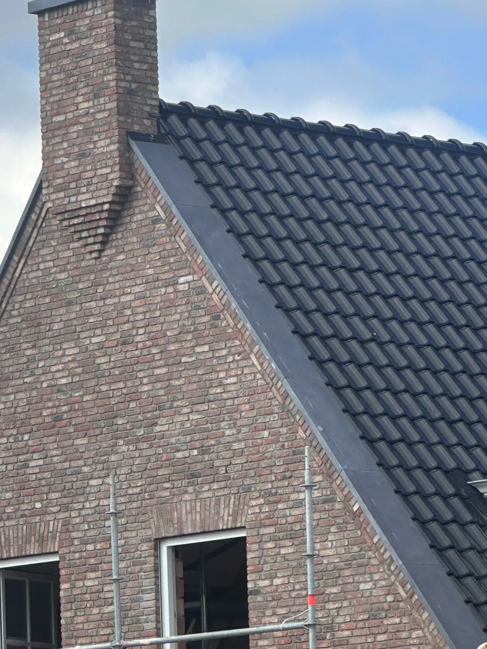 Een gevel van een huis met een schoorsteen, bakstenen muur en een pannendak. Een raam onder de muur en een stalen steiger voor herstelwerkzaamheden.