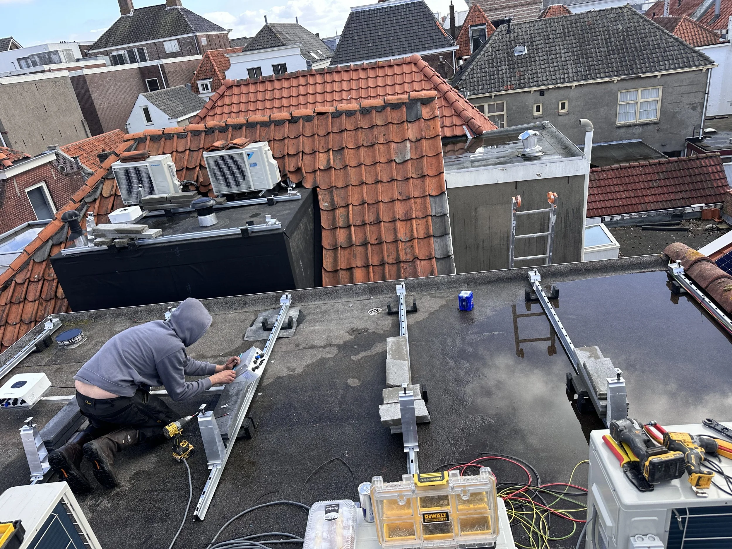 Een technicus installeert solar panelen op een plat dak van een gebouw. Op de achtergrond zijn meerdere huizen met schuine daken zichtbaar.