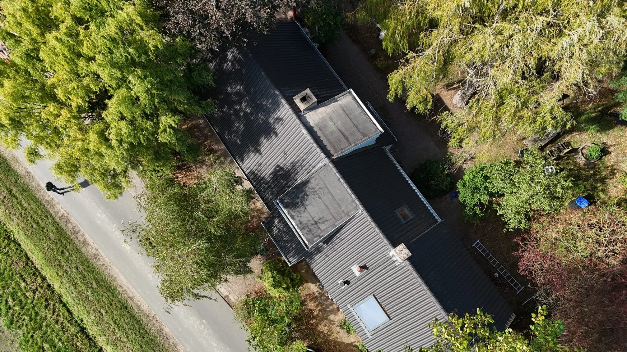 Luchtopname van een huis met een zwart metalen, net gerenoveerd dak omgeven door groene bomen en een weg aan de linkerkant.