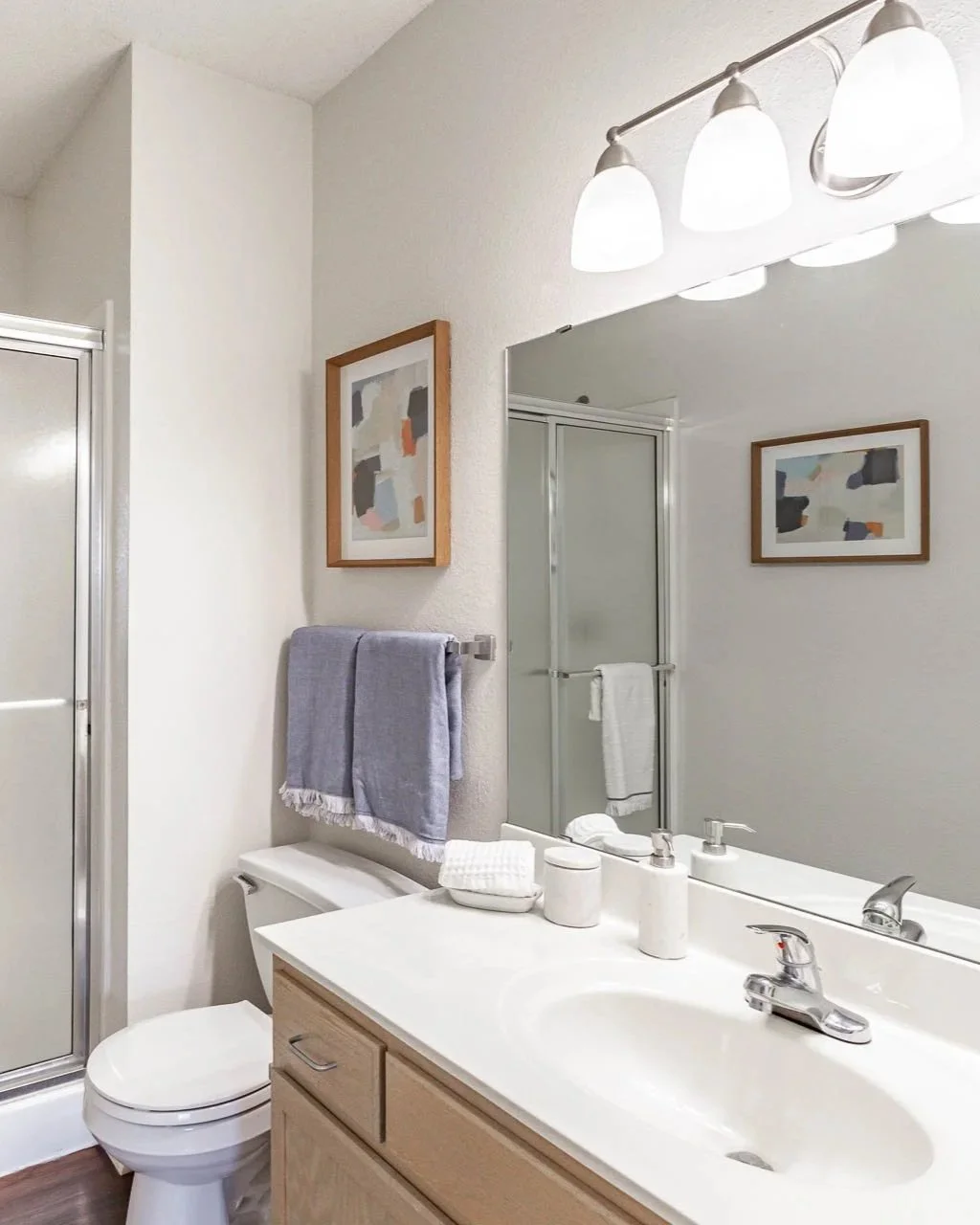 Bathroom with a white sink vanity, a large mirror, framed abstract art, a towel rack with towels, a toilet, and a shower with frosted glass door.