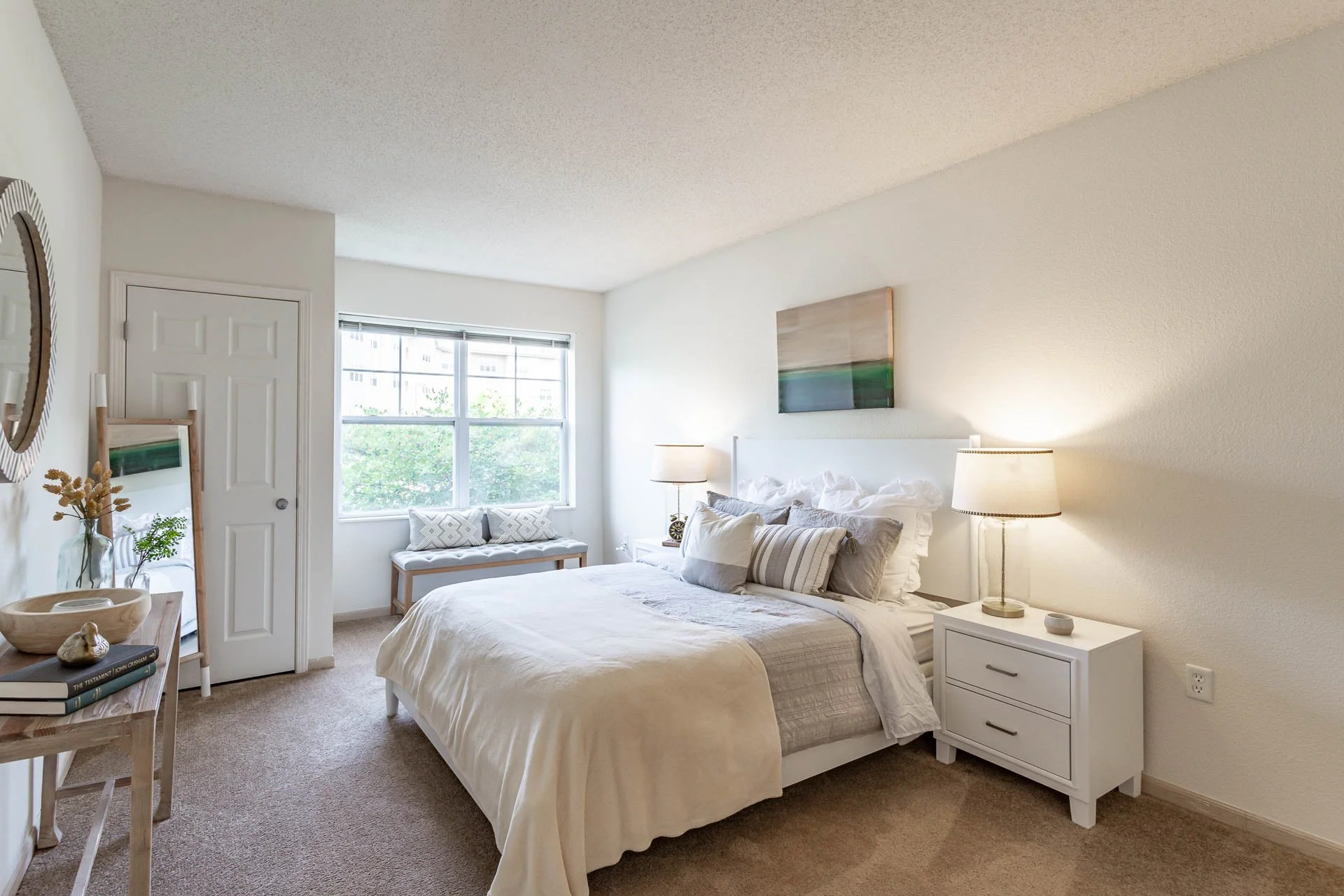 A bedroom with a white bed, pillows, bedside lamps, a window with a bench seat, and minimalist decor.