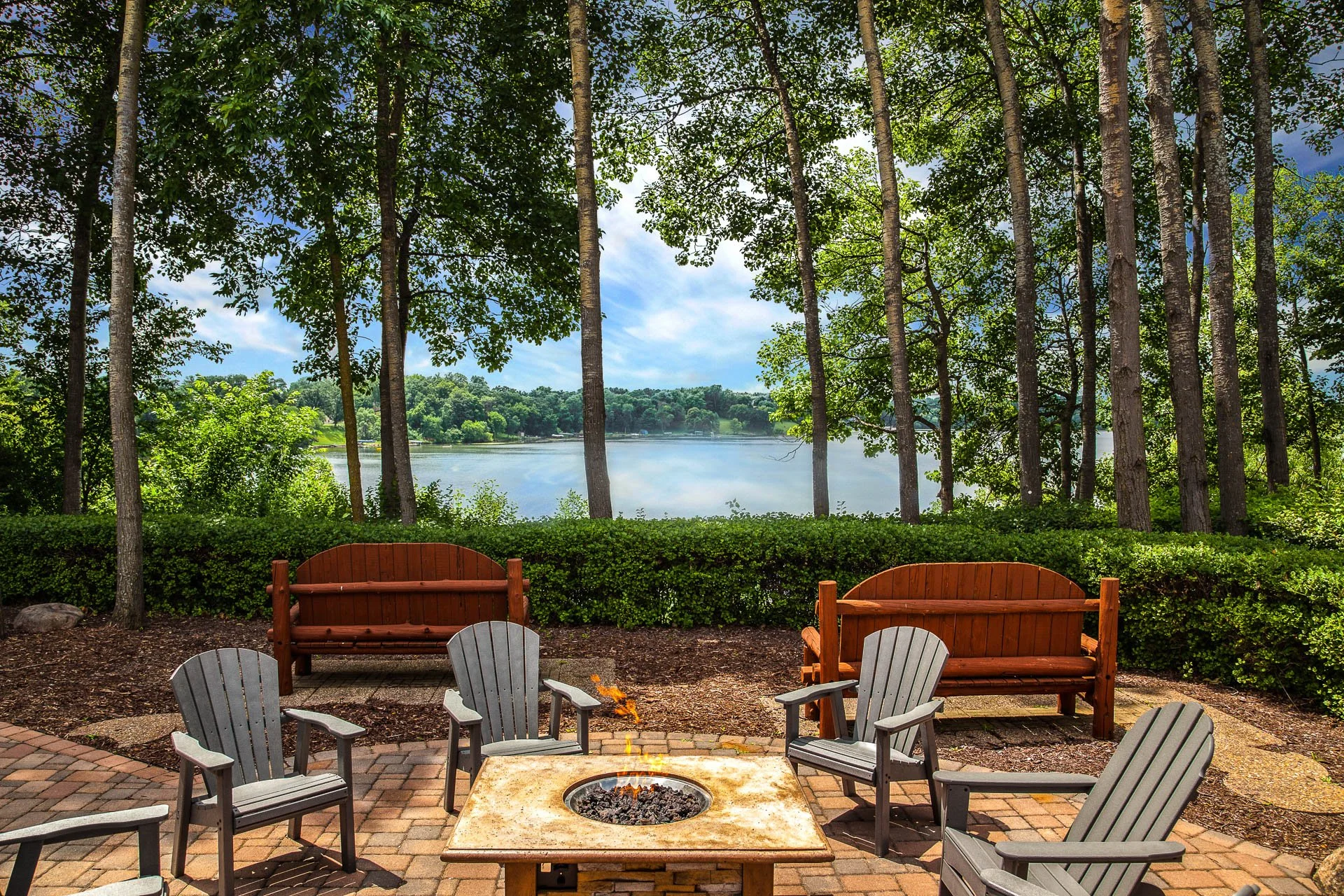 Outdoor patio with a firepit and Adirondack chairs, overlooking a lake and surrounded by trees.
