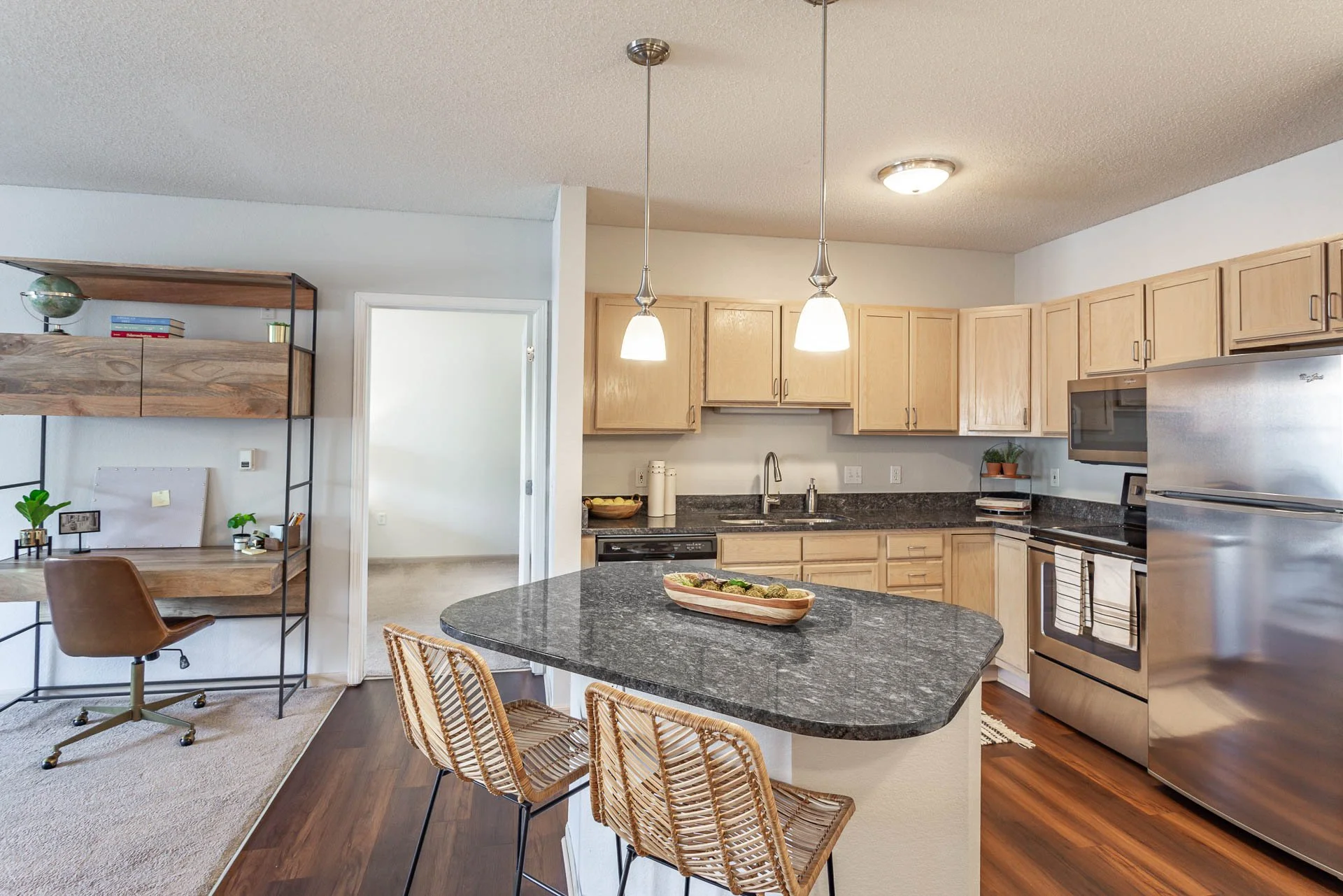 Kitchen with light wood cabinets, stainless steel refrigerator, dishwasher, and oven, with a black granite countertop, a small kitchen island with bar stools, and hanging pendant lights.