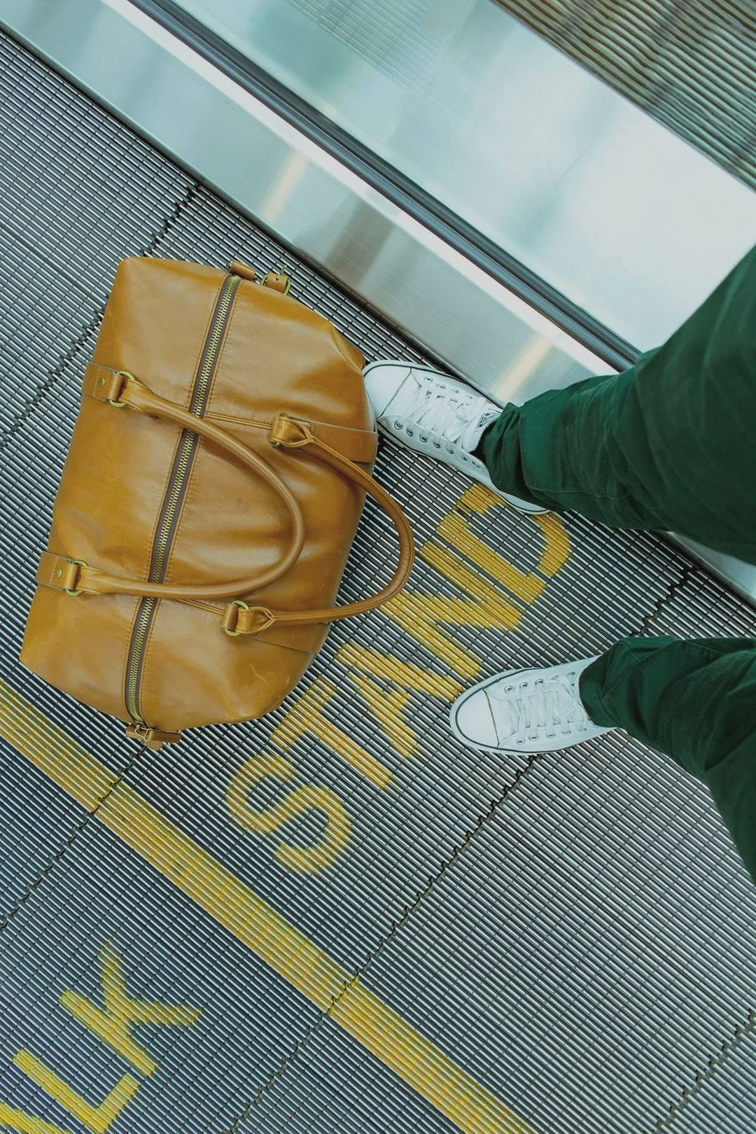 Person wearing white sneakers and green pants standing next to a brown leather duffel bag on a metal grid floor at an airport gate, with yellow markings.