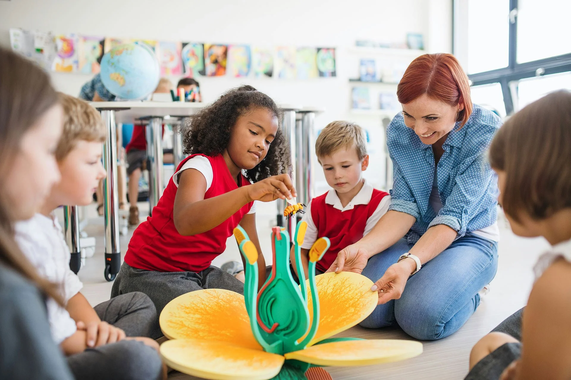 A teacher and young students sitting on the floor around a colorful educational model of a flower, engaging in a classroom activity.