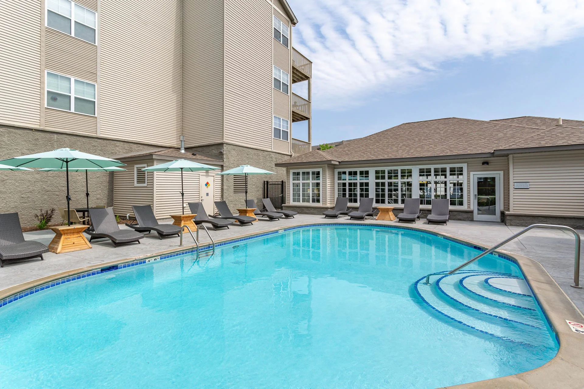 Apartment complex outdoor swimming pool area with lounge chairs, umbrellas, and a surrounding building with windows and balconies under a partly cloudy sky.