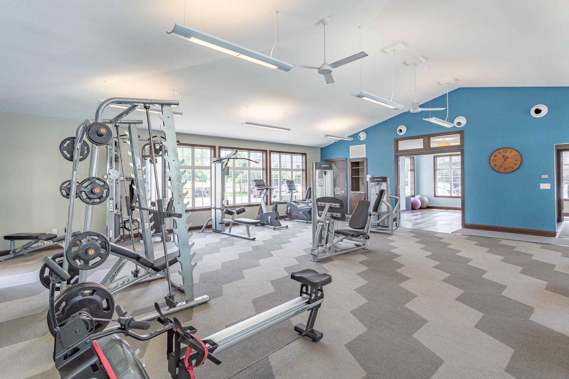 Empty gym with various exercise equipment, large windows, and blue accent wall.