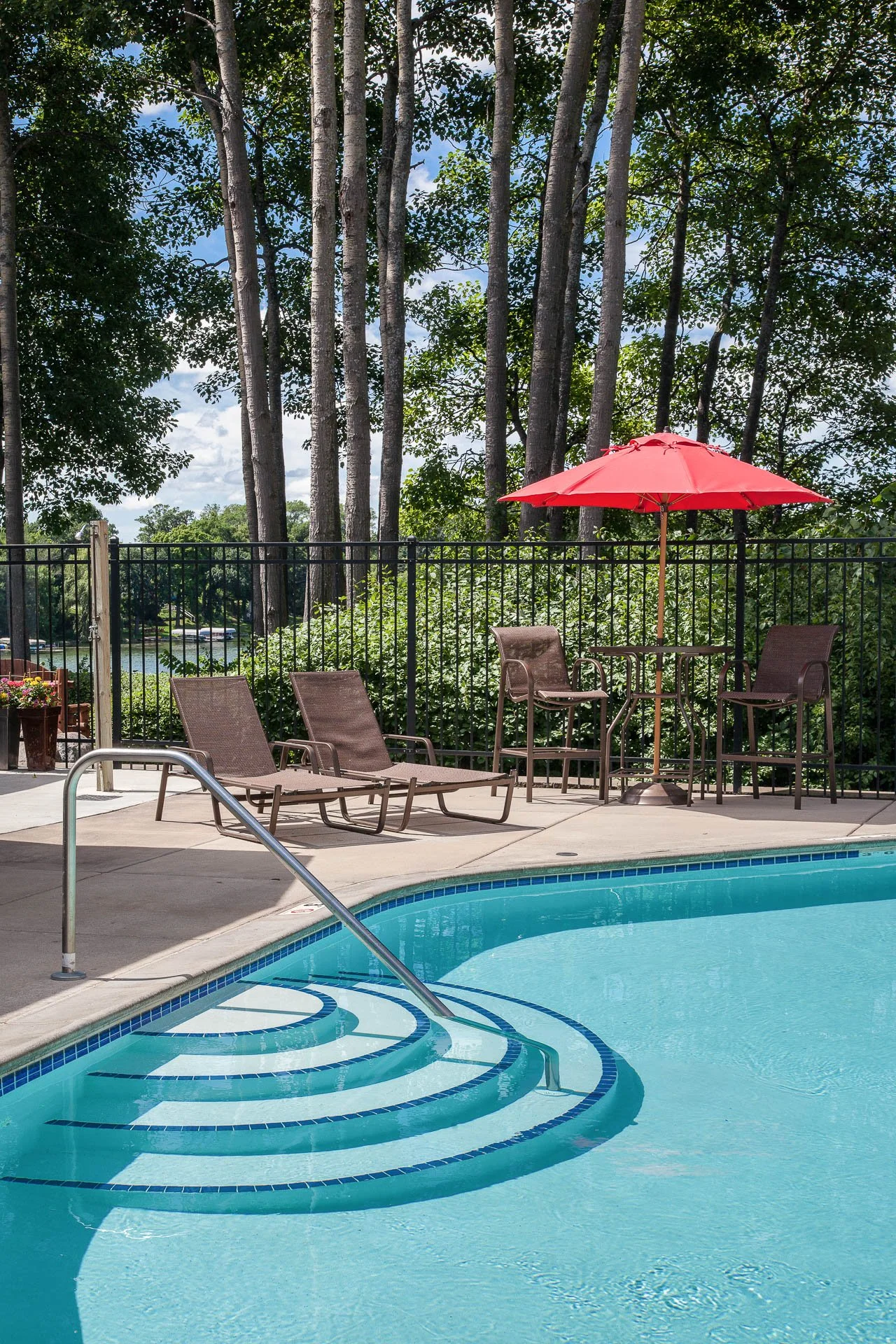 Poolside area with lounge chairs, a table with an umbrella, and a black fence with trees and a body of water in the background.