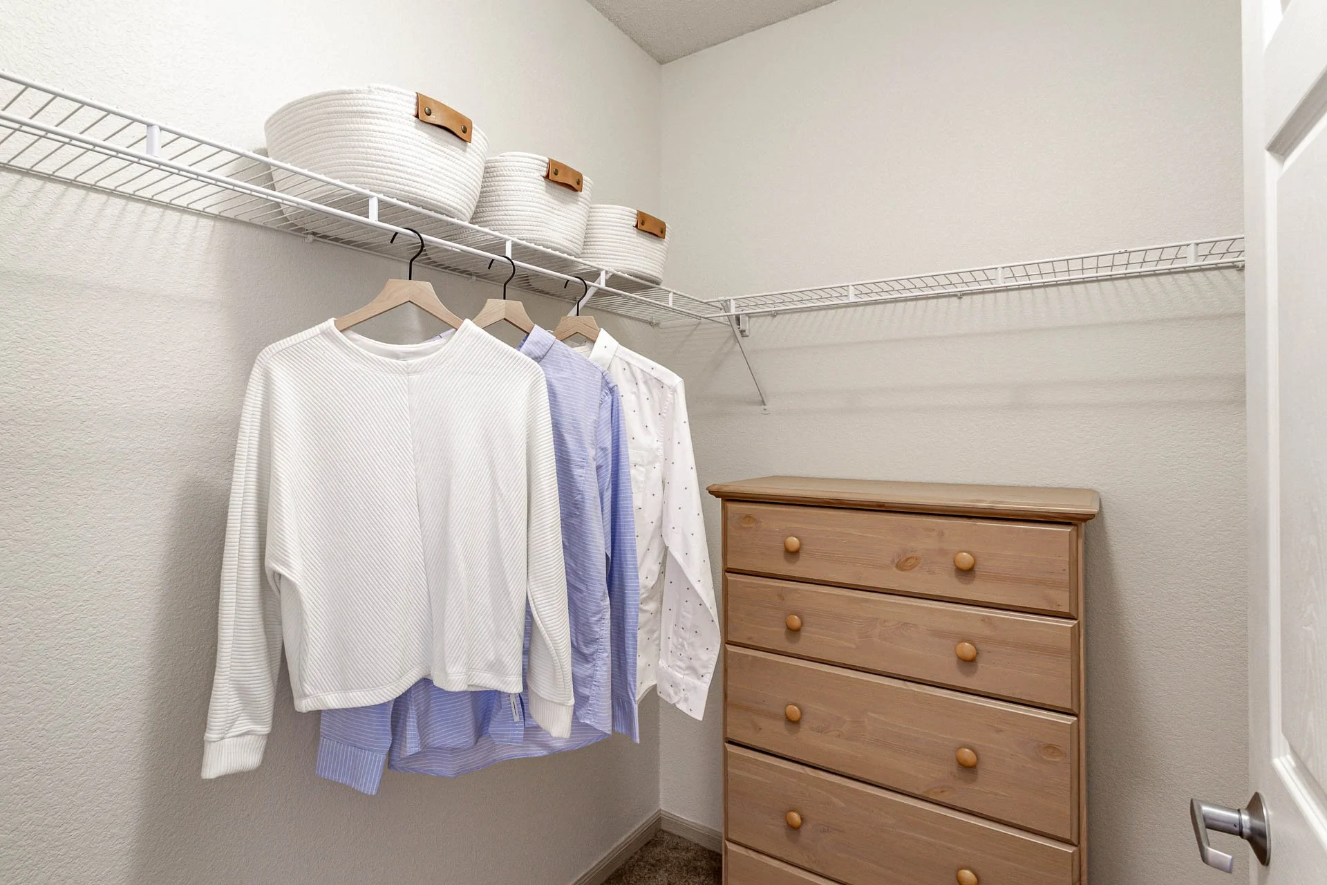 Empty walk-in closet with hanging clothes, woven baskets on a wire shelf, and a wooden dresser.