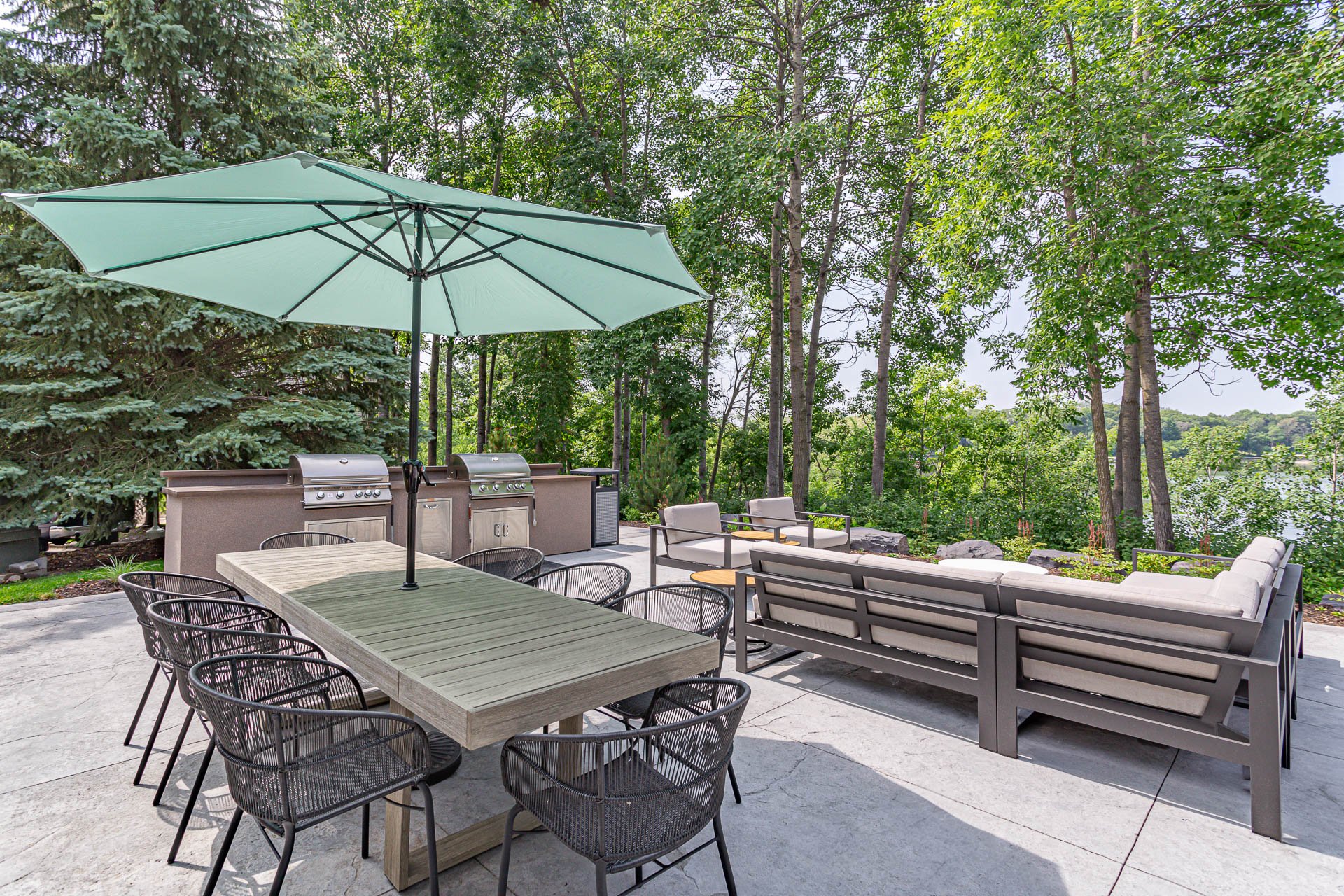 Outdoor patio area with a dining table, chairs, a large umbrella, a sitting area, and a built-in grill, surrounded by lush green trees.