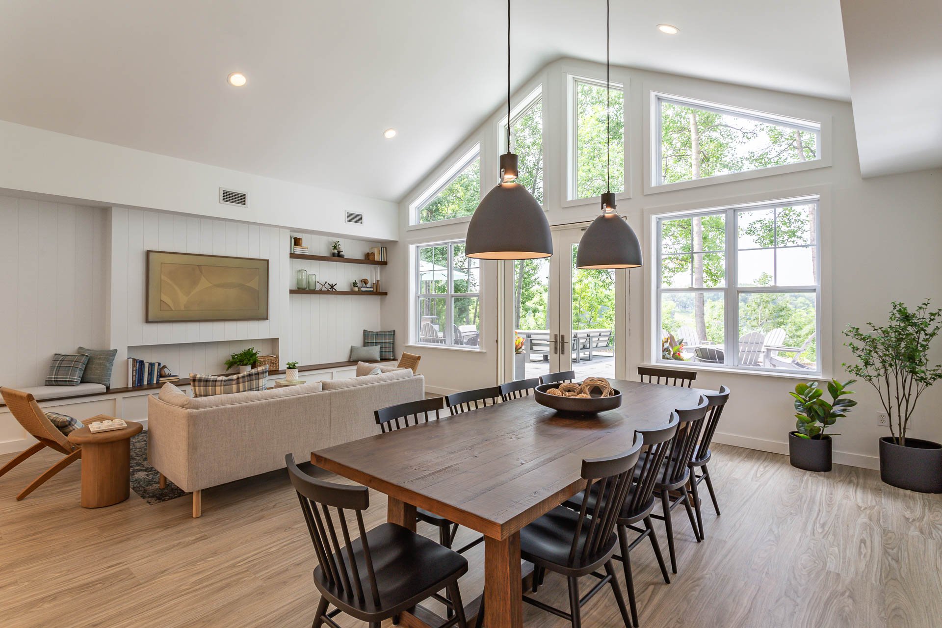 Open-concept living and dining room with large windows, beige sofa, wooden dining table, black chairs, and houseplants.