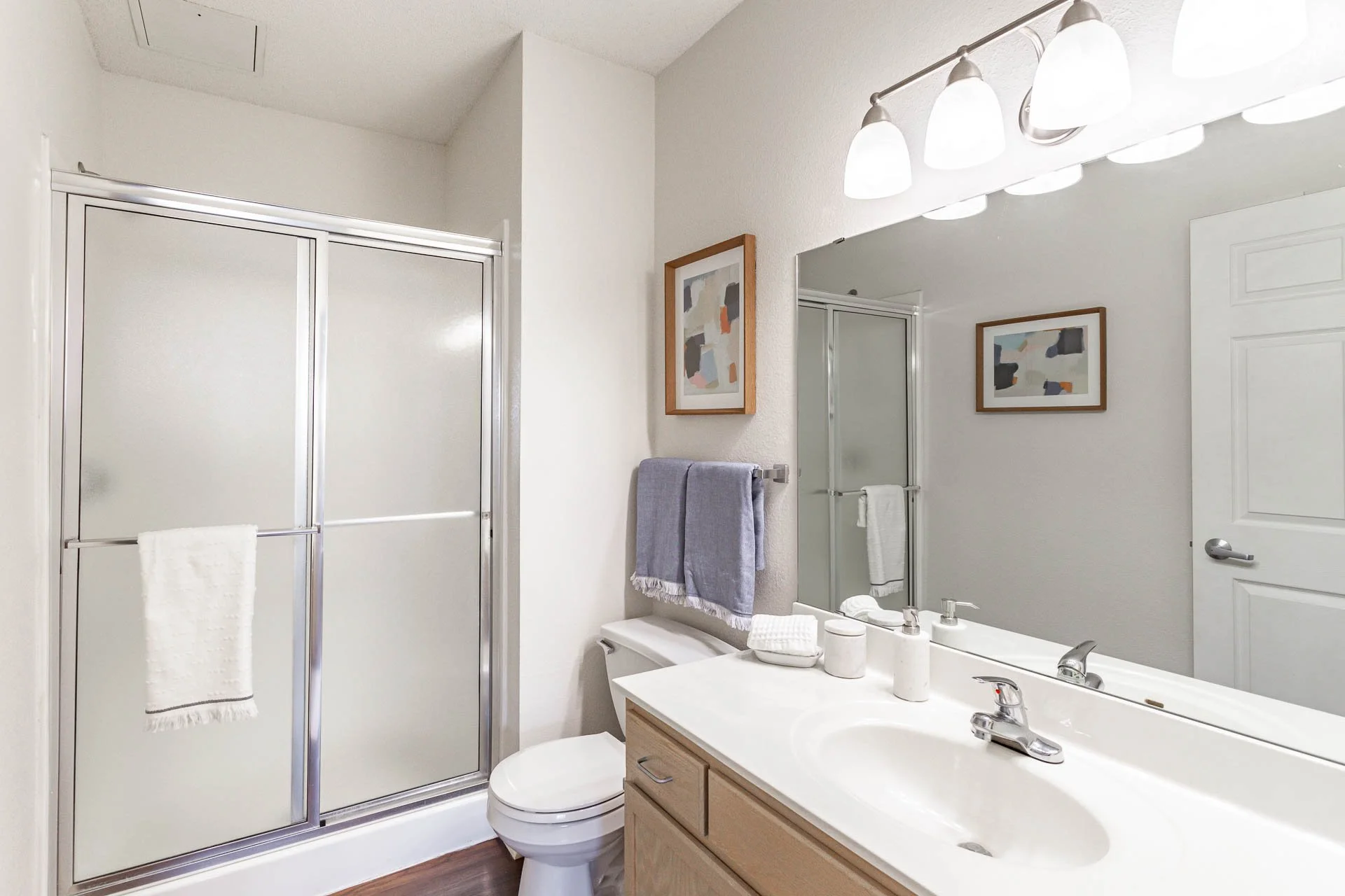 A clean bathroom with a shower stall with frosted glass door, a toilet, a wooden vanity with a white countertop, a large mirror, a modern sink and faucet, a shelf with decorative items, and framed abstract art on the wall.