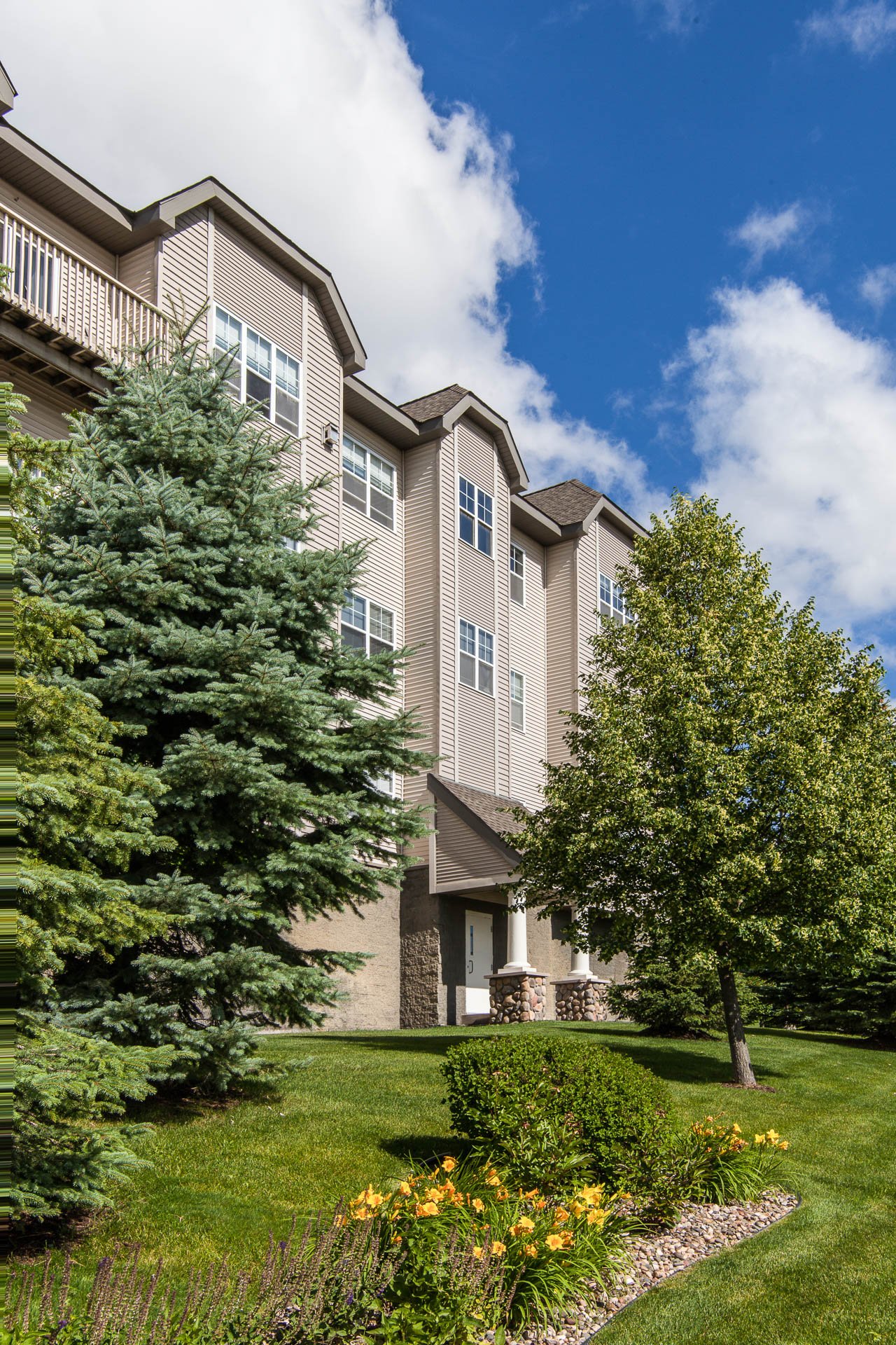 A multi-story beige apartment building with white-framed windows, surrounded by lush green trees, a well-maintained lawn, and colorful flowers, under a partly cloudy blue sky.