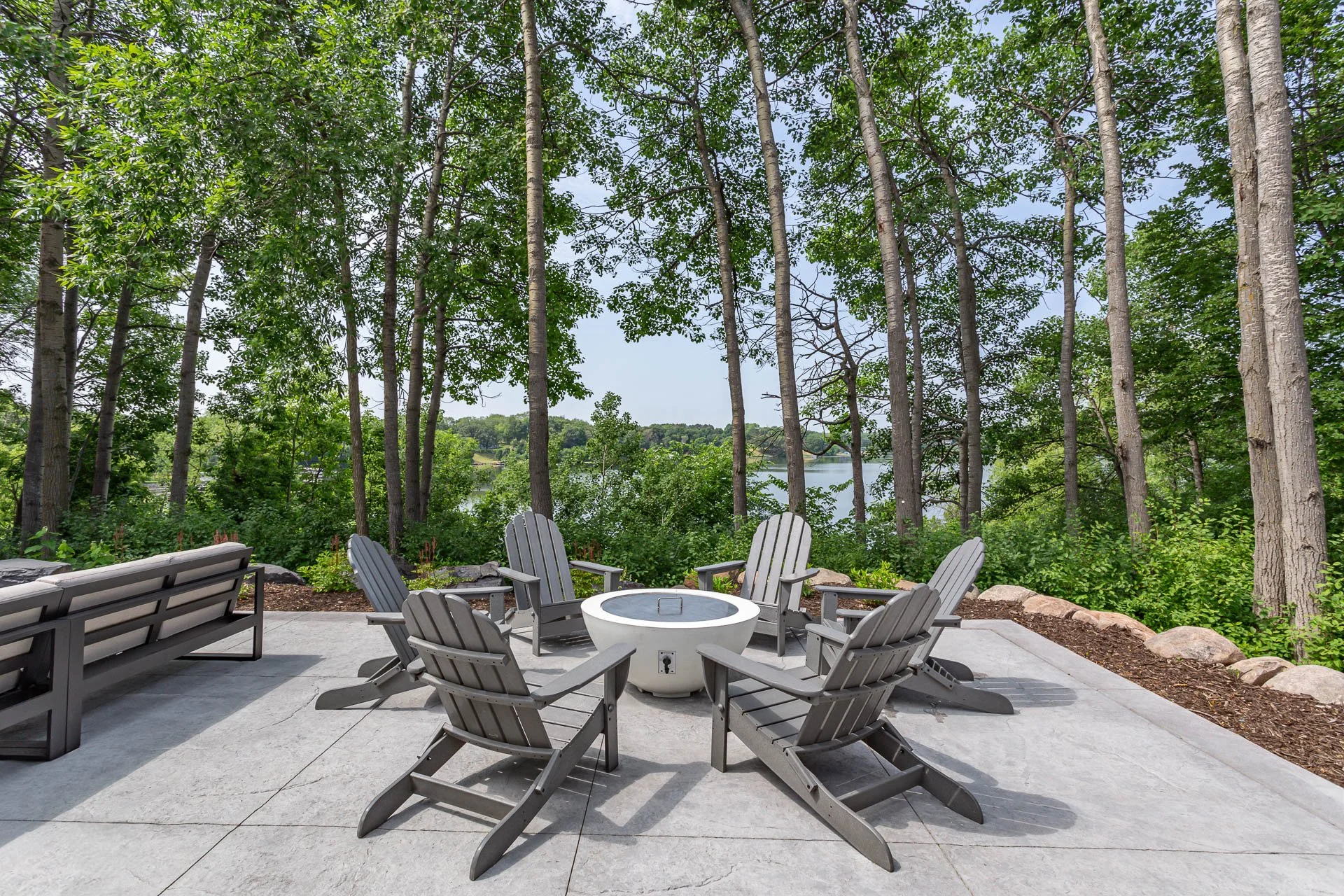 Outdoor patio with six gray Adirondack chairs arranged in a circle around a fire pit, with a view of a lake and trees in the background.