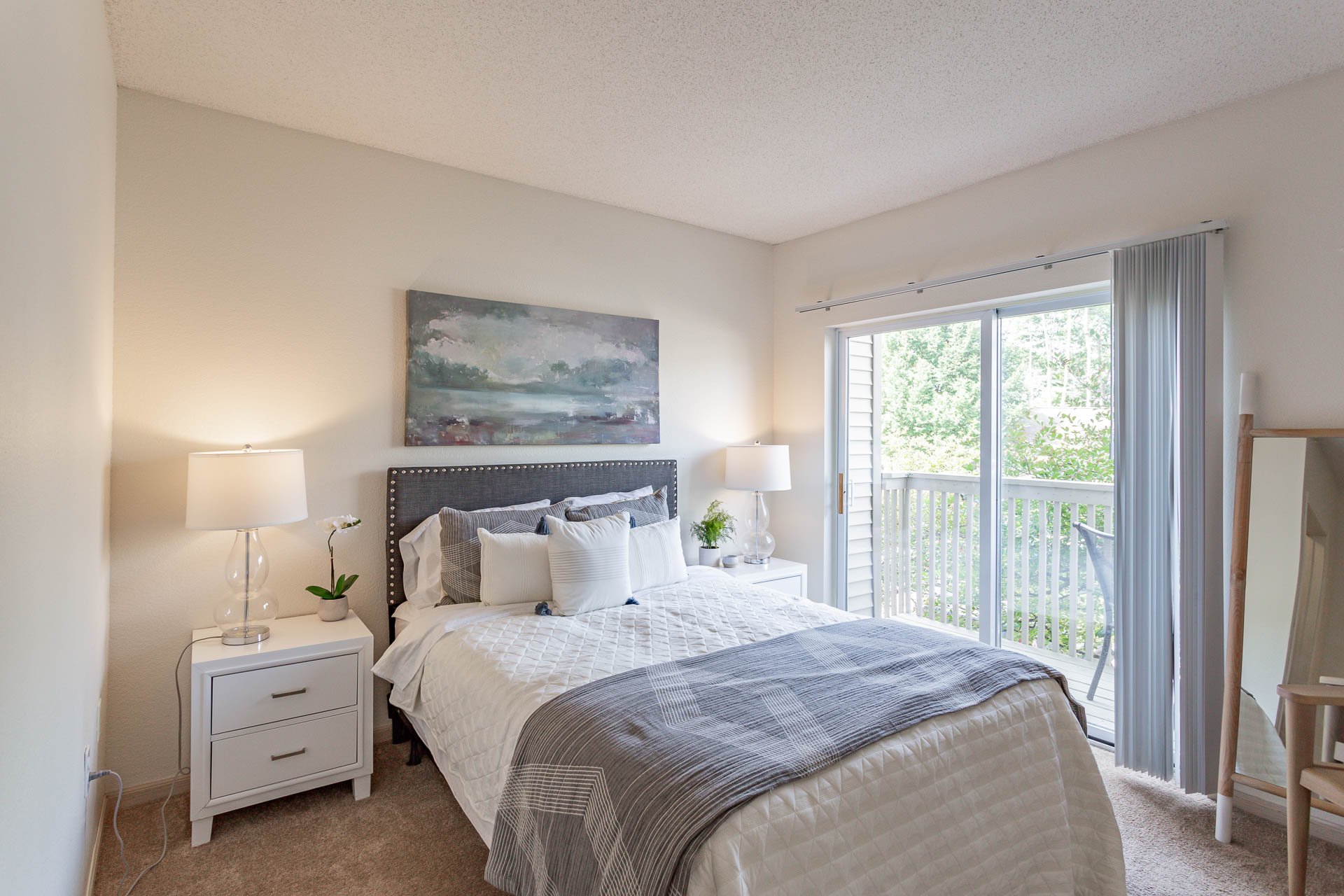 Bright bedroom with a bed, nightstands, lamps, and a sliding glass door leading to a balcony with trees outside.