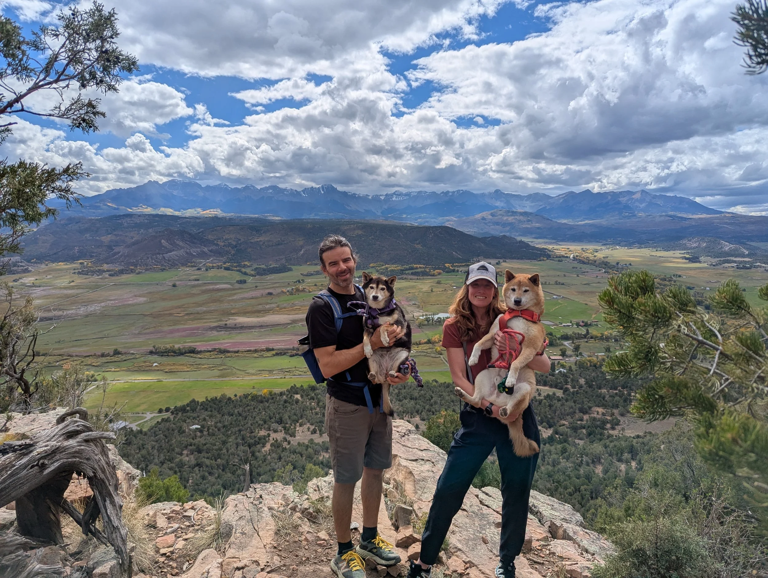 A man and a woman holding two dogs pose on a rocky trail with a valley, mountains, and partly cloudy sky in the background.