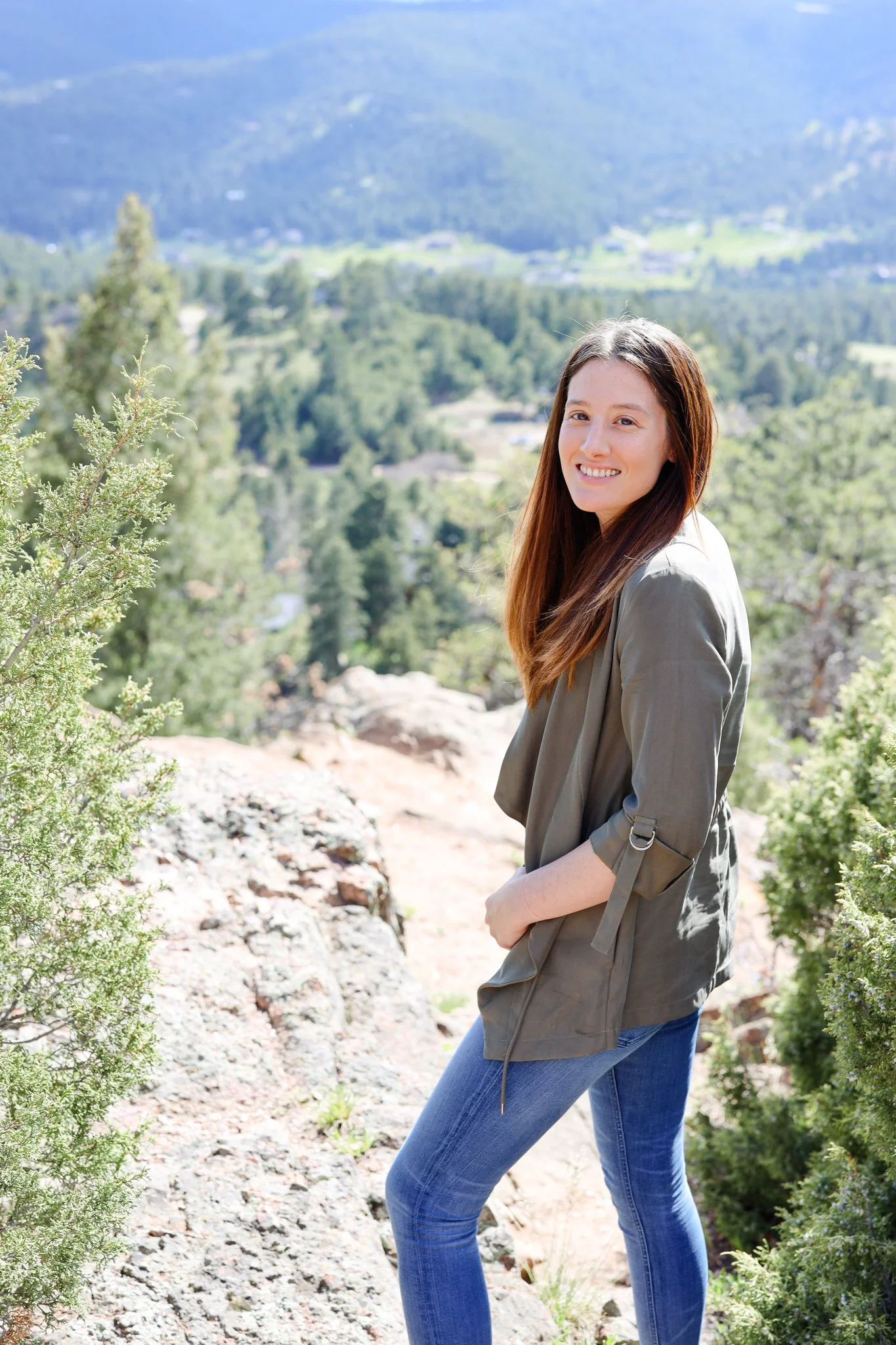 A young woman with long brown hair smiling outdoors on a sunny day, wearing a gray jacket and jeans, with a scenic mountain and forest background.