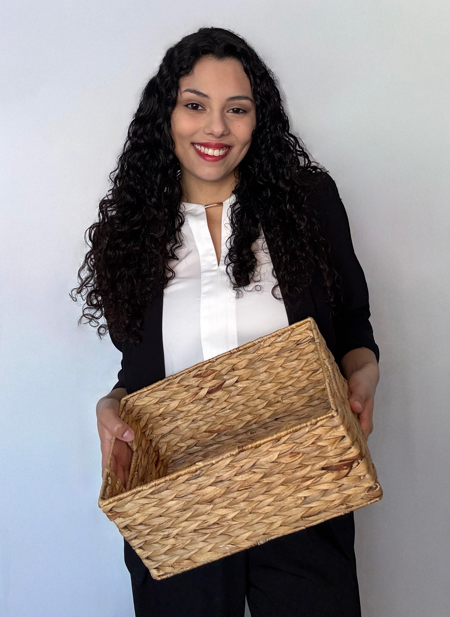A woman with long curly black hair smiling and holding a woven basket, dressed in a black blazer over a white blouse, standing against a plain white wall.