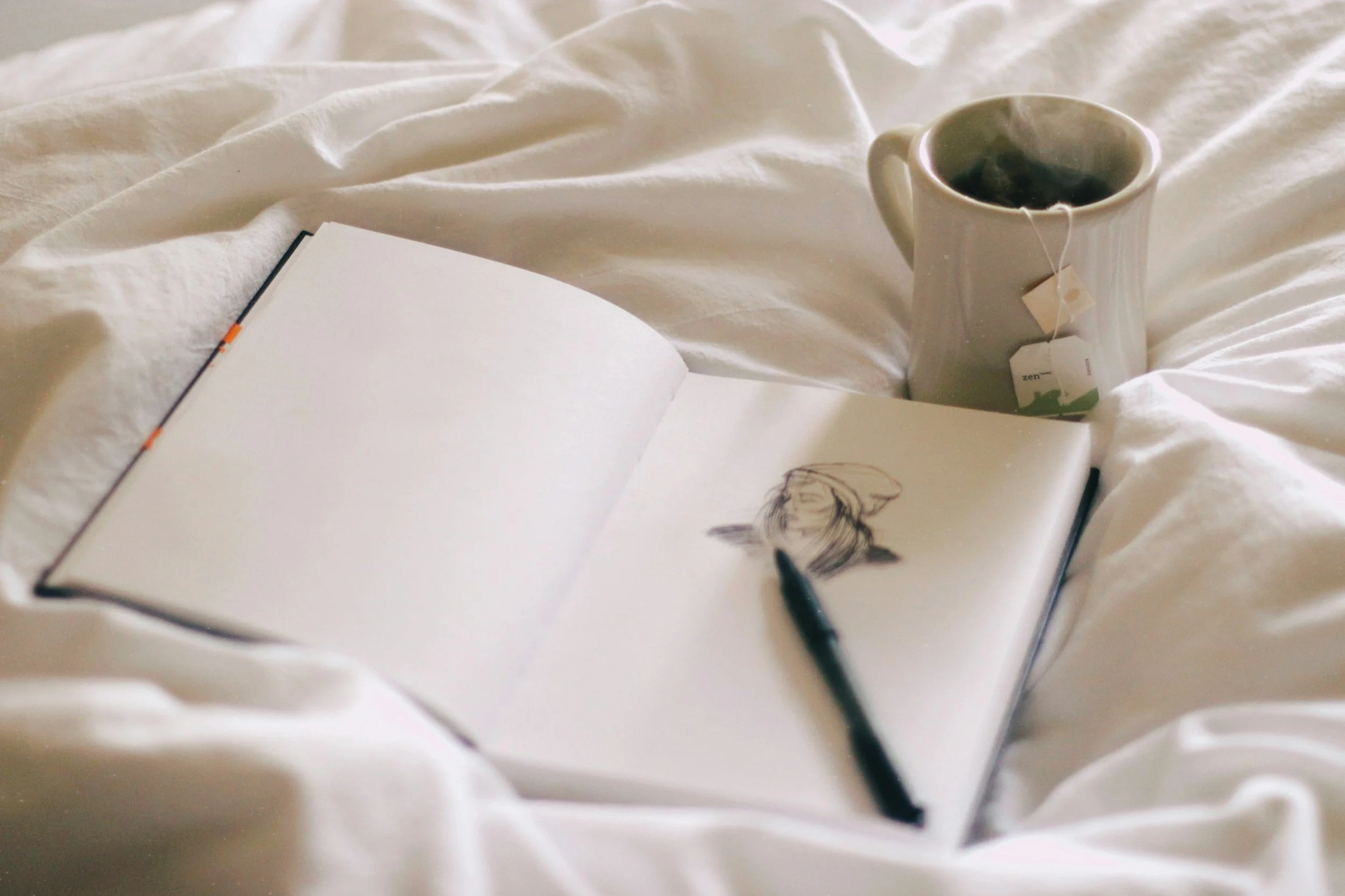 An open sketchbook with a black pen on a white bed, along with a ceramic mug of tea with a tea bag, and a simple pencil sketch of a woman's face on the right page.