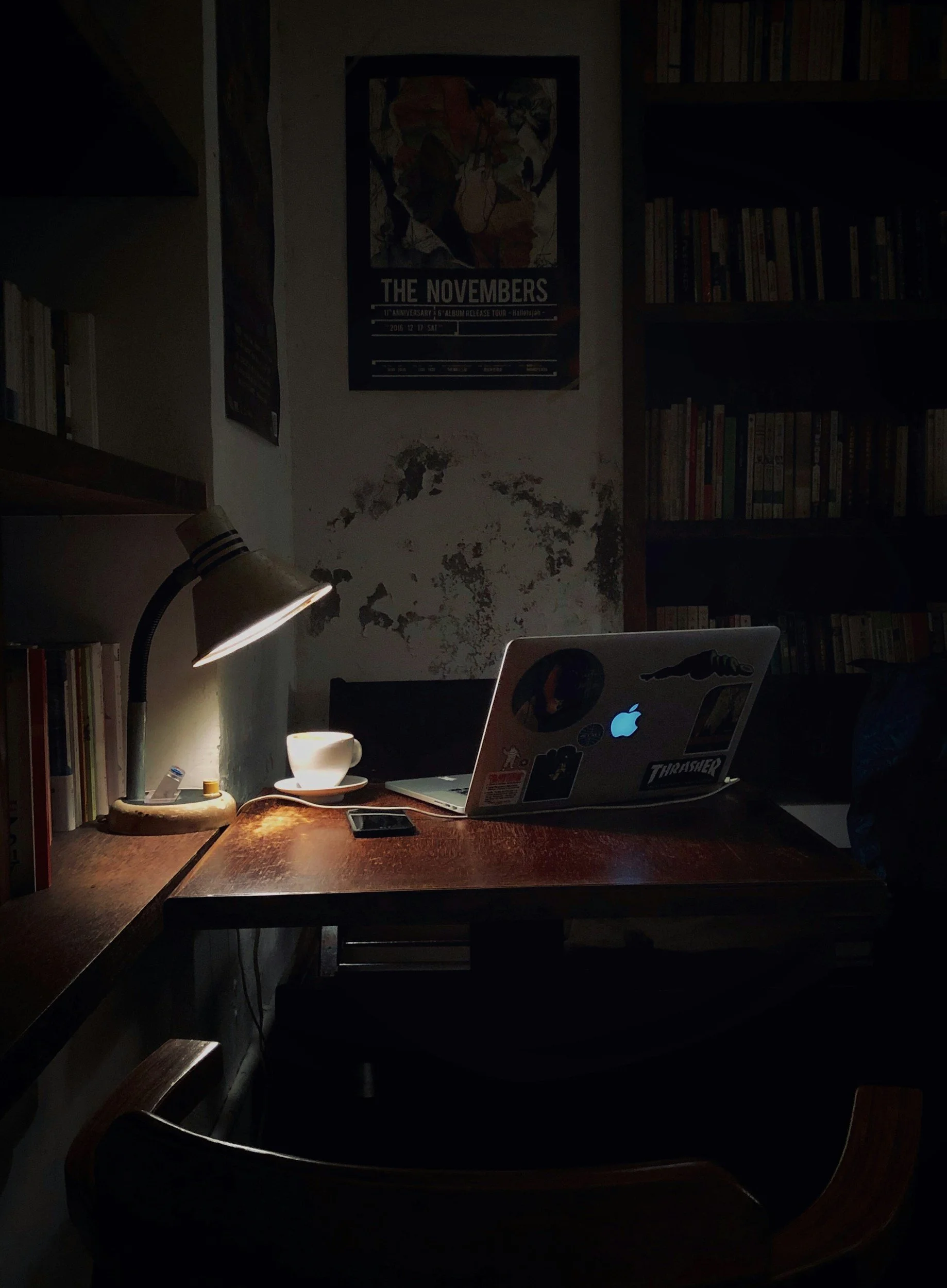 A dimly lit workspace with a wooden desk, a laptop with stickers, a smartphone, a coffee cup on a saucer, and a desk lamp illuminating the area. There are bookshelves and posters on the wall in the background.