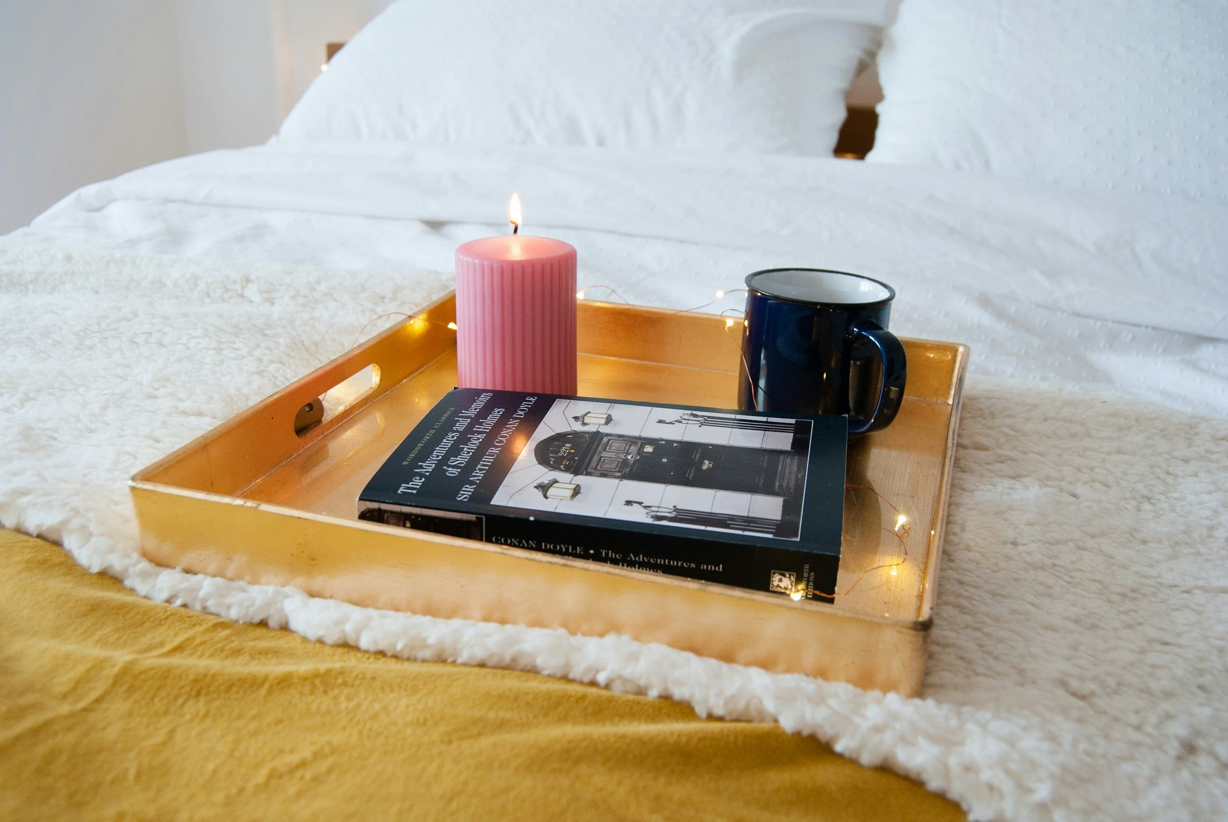 A wooden tray on a bed holding a pink candle, a navy blue mug, a book titled 'The Adventures and Secrets of Sherlock Holmes' by Conan Doyle, and string fairy lights, with white bedding and pillows in the background.
