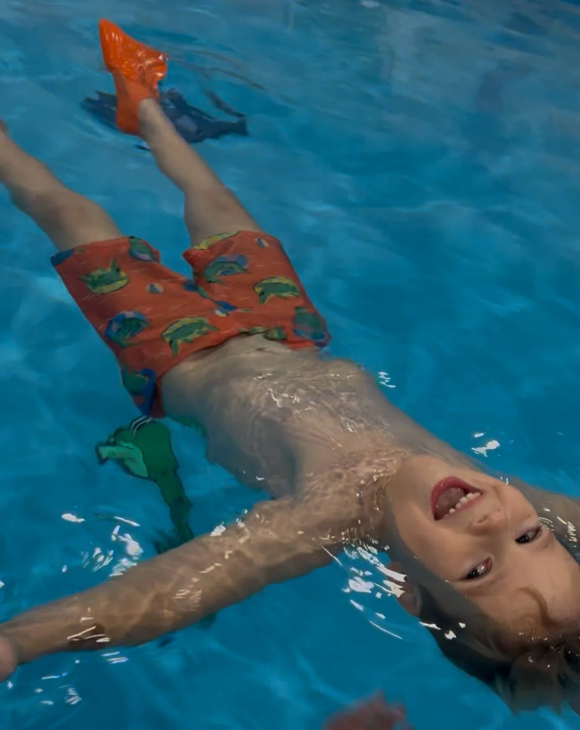 Child swimming in a pool, smiling, wearing orange swim shorts with colorful fish pattern and orange swim shoes.