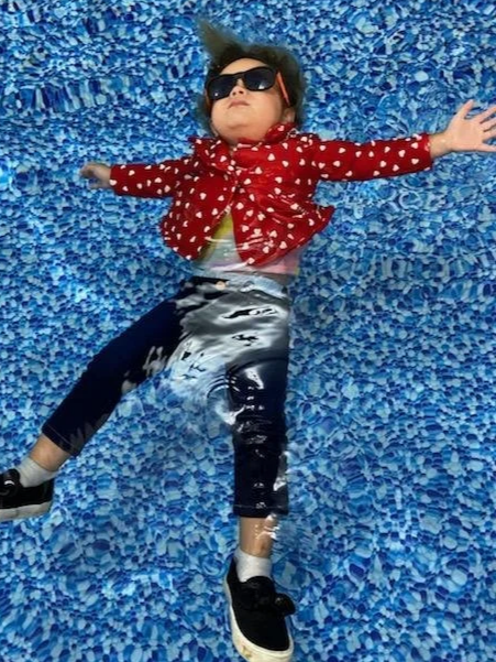 Child laying on a blue textured surface, wearing sunglasses, a red polka dot jacket, and black and white pants.