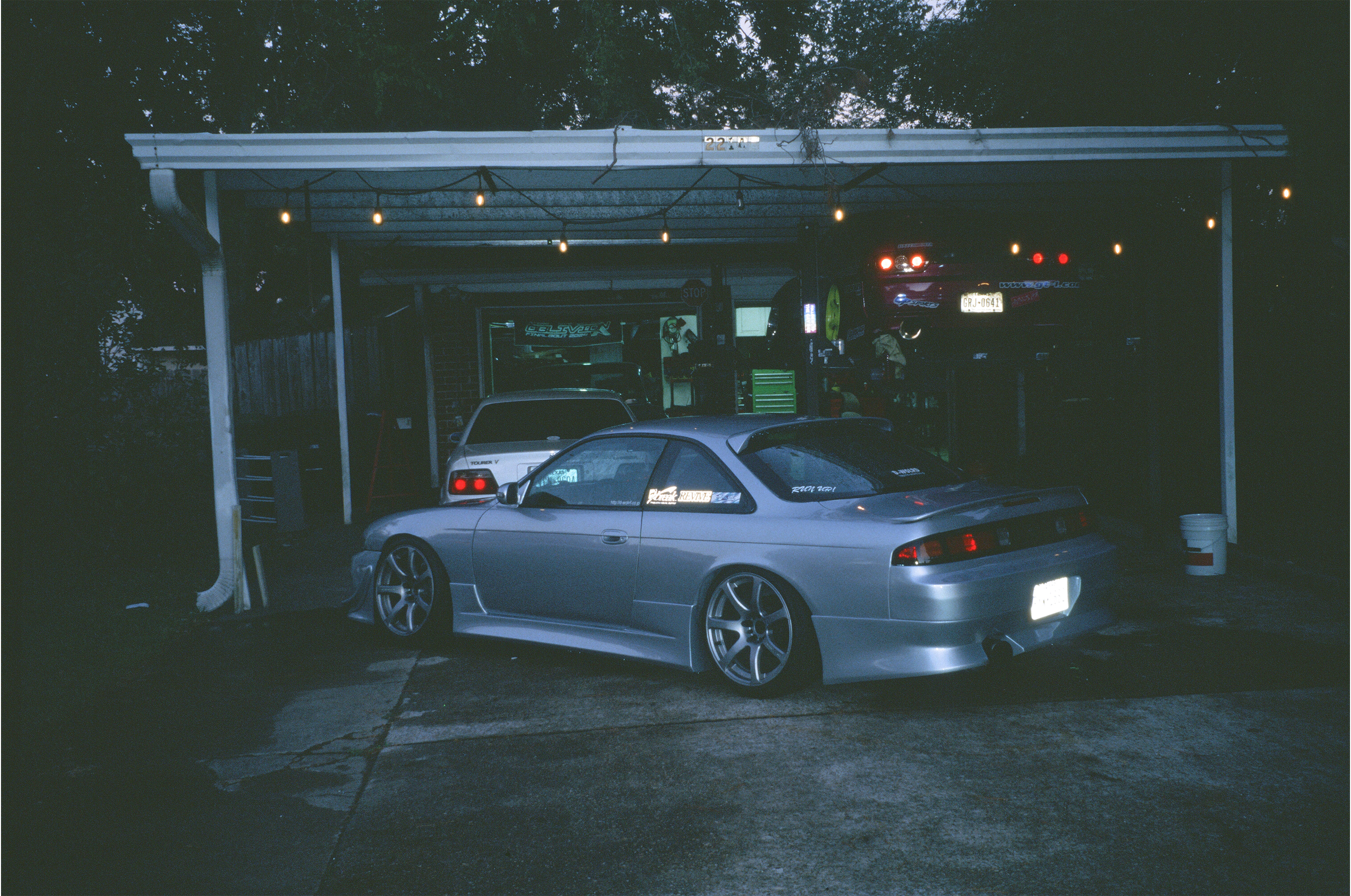 A silver sports car is parked in front of a garage with a car lift and tools. There are string lights hanging above the area, and another car is elevated on the lift inside the garage. The garage has a brick wall, and some trees are visible in the background.