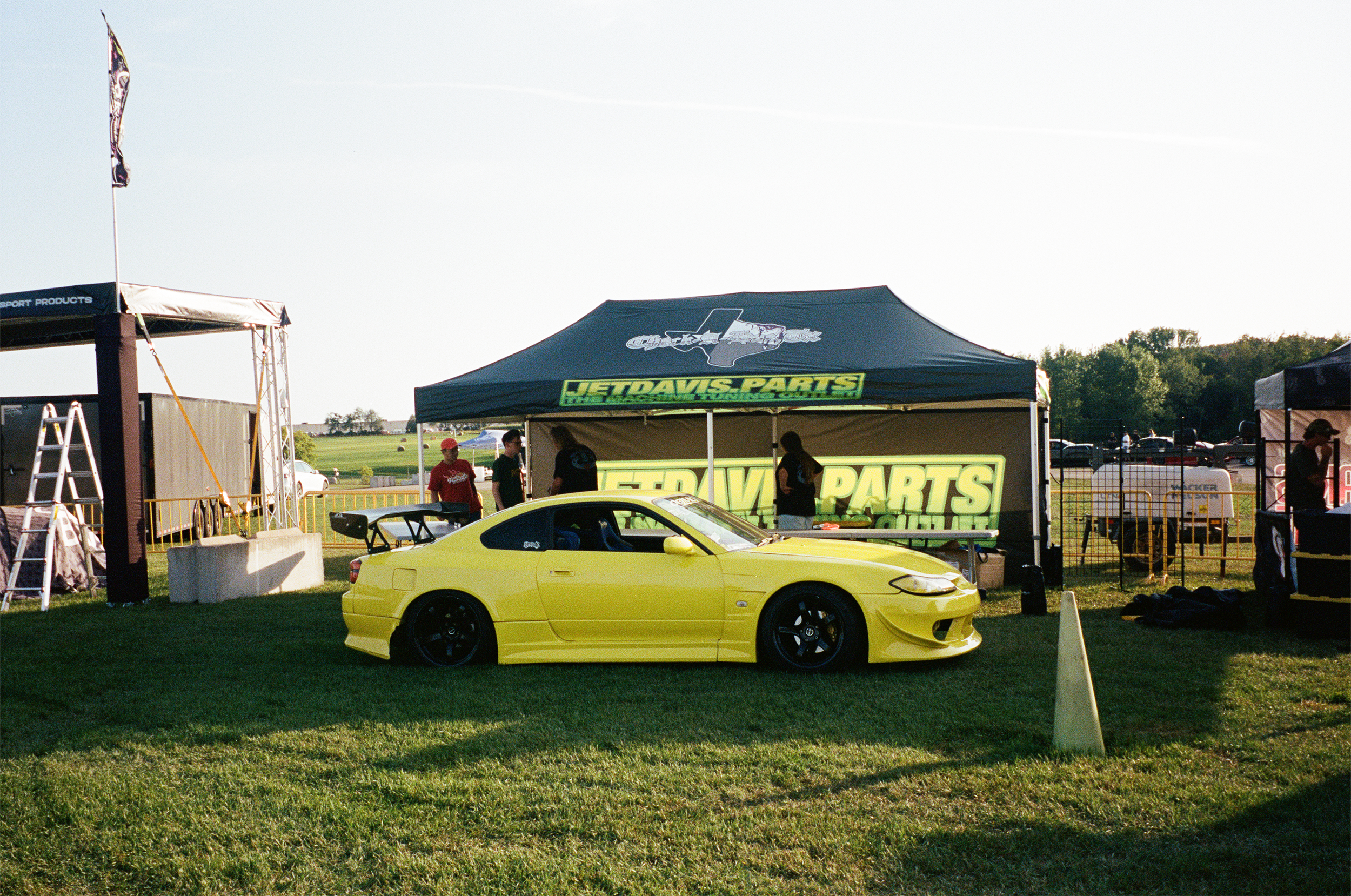 Yellow sports car parked on grass at an outdoor event, with tents and people in the background, including a tent with 'JetDavies Parts' branding.
