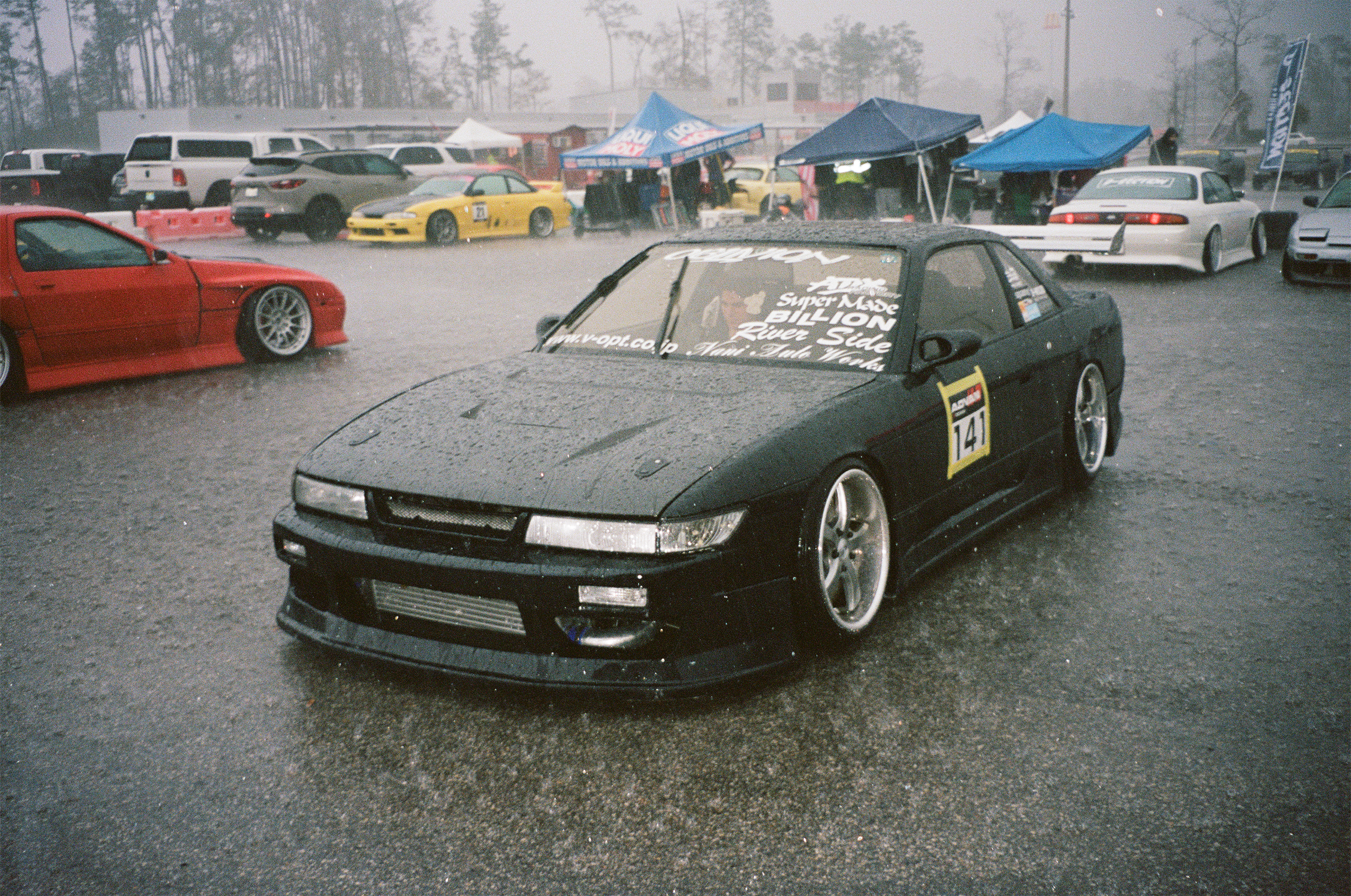 Black race car with yellow number 41 on the side, parked on a wet gravel surface during a rainy day at a car event, with tents and other vehicles in the background.