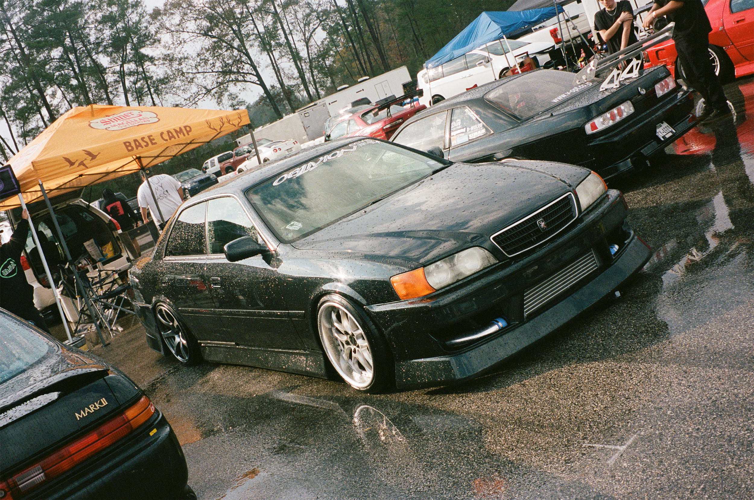 Black modified JZX100 with lowered stance and aftermarket wheels parked at a car meet, wet from rain, with other cars and a yellow tent in the background.