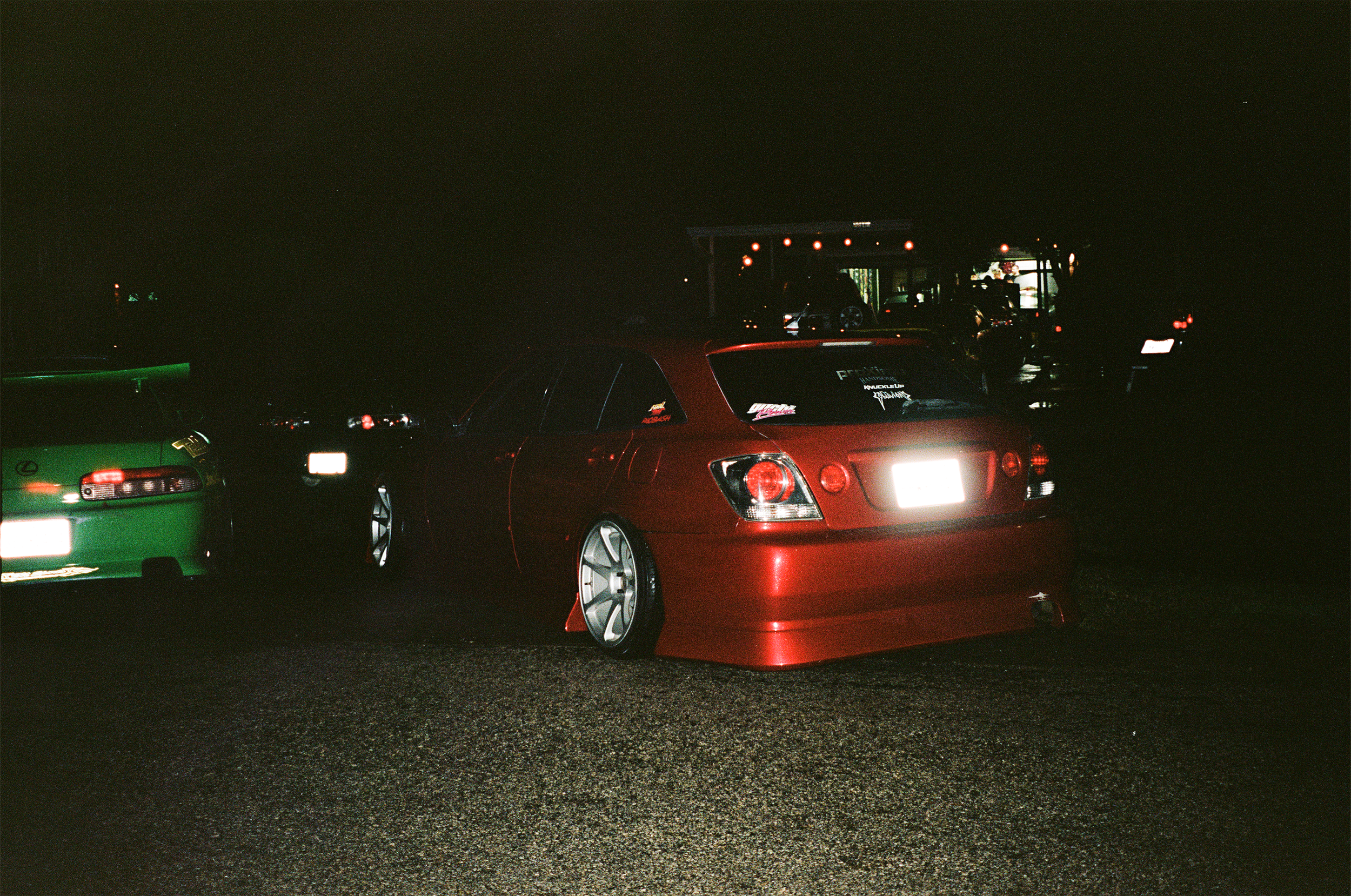 Nighttime scene of a red customized car with lowered suspension, large silver wheels, and a rear spoiler, parked on a dark street with other cars in the background.
