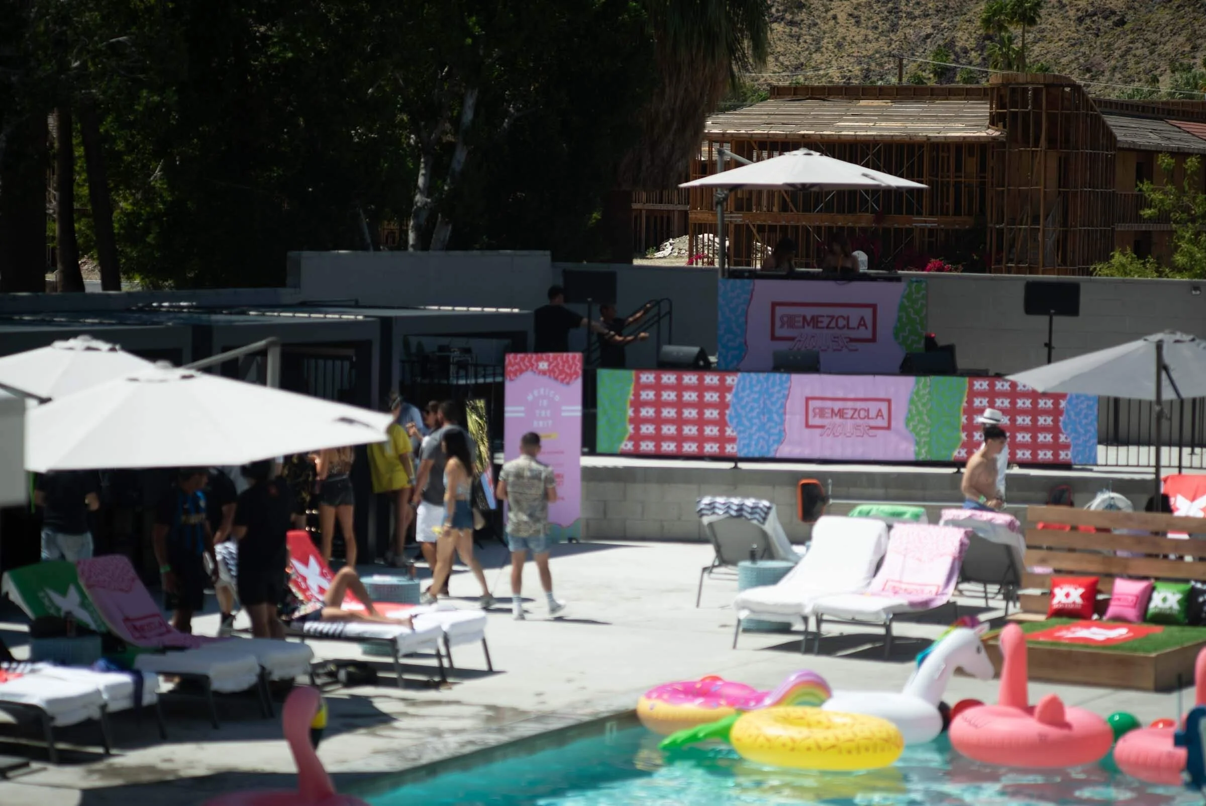 Escenario al aire libre con personas en una piscina y como fondo una pantalla gigante con el texto 'Remezcla House', sombrillas blancas, sillas de playa, flotadores y gente disfrutando del día junto a estructuras en construcción al fondo.