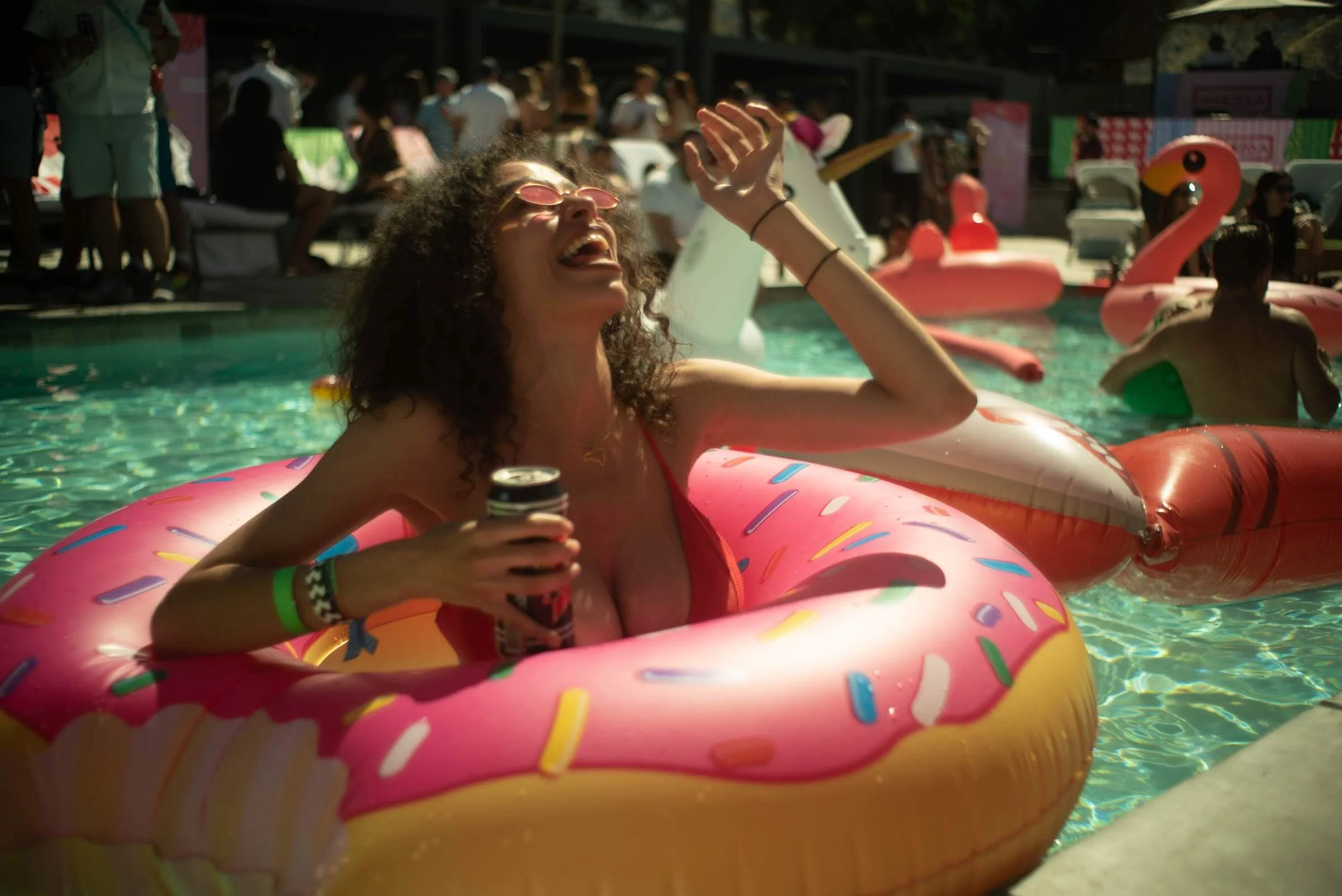 Mujer en una piscina con un inflable de rosquilla con glaseado rosa y chispas de colores, sosteniendo una lata, disfrutando de una fiesta con muchas personas y pelotas inflables en el agua y alrededor de la piscina.