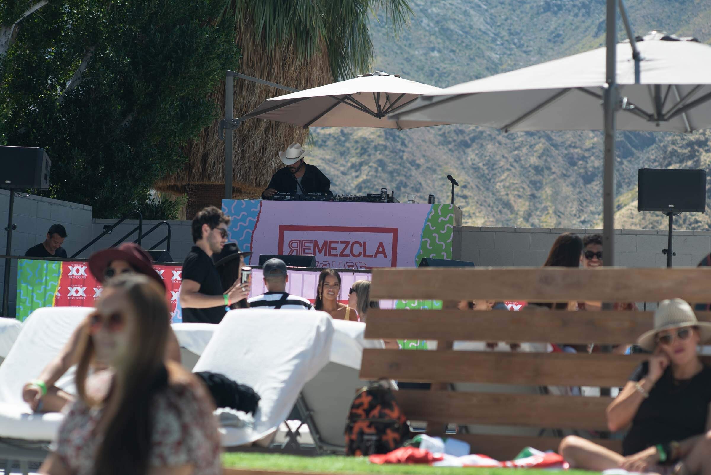 Escenario al aire libre con personas disfrutando un evento, con DJ en una cabina bajo sombrillas y montañas de fondo.