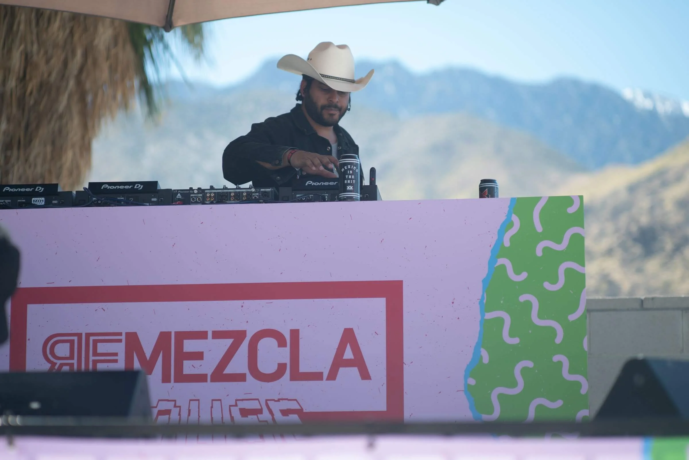 Un DJ con sombrero de vaquero mezclando música en un escenario al aire libre con montañas al fondo. Parte de un cartel en primer plano con la palabra 'Mezcla' en letras grandes.